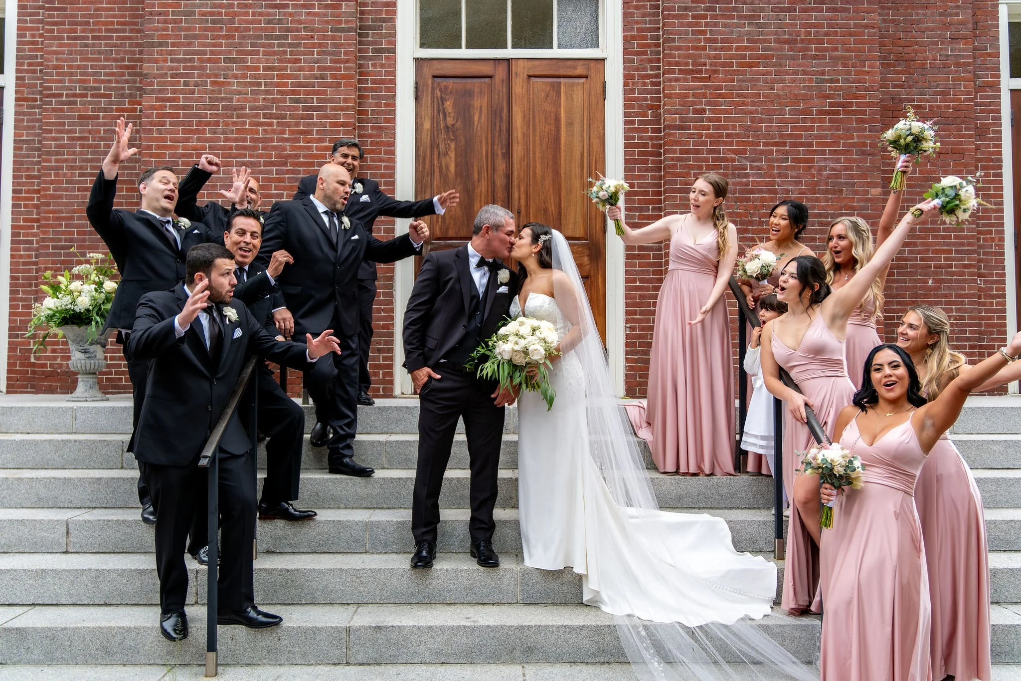 Nicole and Alexey kissing on the church steps surrounded by their wedding party at Holy Trinity Greek Orthodox Church Portland Maine summer wedding photography VMG Productions