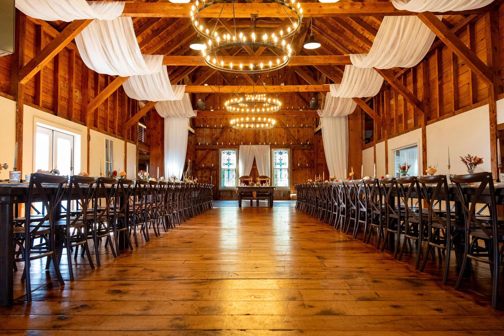 Wide view of the Boothbay Playhouse barn set with long farm tables, wooden chairs, white draping, and chandeliers at Boothbay Playhouse in Boothbay Maine fall wedding photography VMG Productions