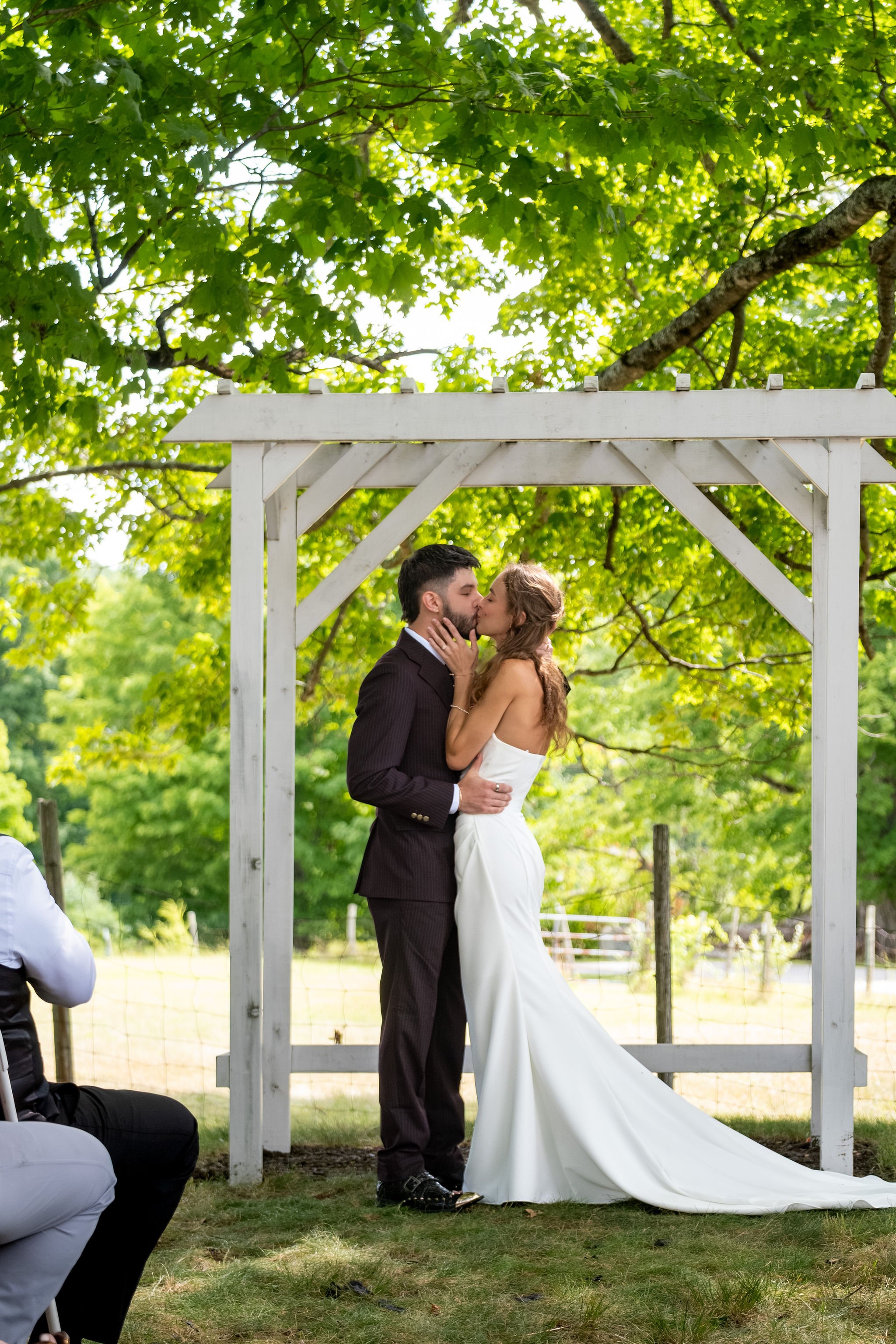 Cassidy and Chase Begin's first kiss as a married couple under the white arbor at Maple Hill Farm Inn and Conference Center in Hallowell Maine summer wedding photography VMG Productions