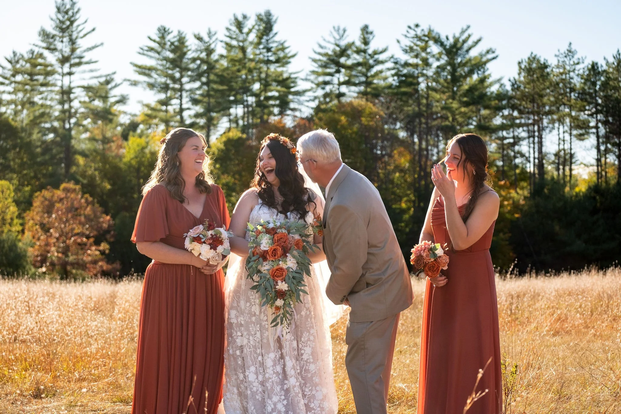 Bride Meagan in floral lace wedding gown and flower crown laughing with father in tan suit during candid moment between groupings with two bridesmaids in terracotta dresses reacting with delight in golden autumn meadow at Watson Homestead Hiram Maine fall 2025 wedding
