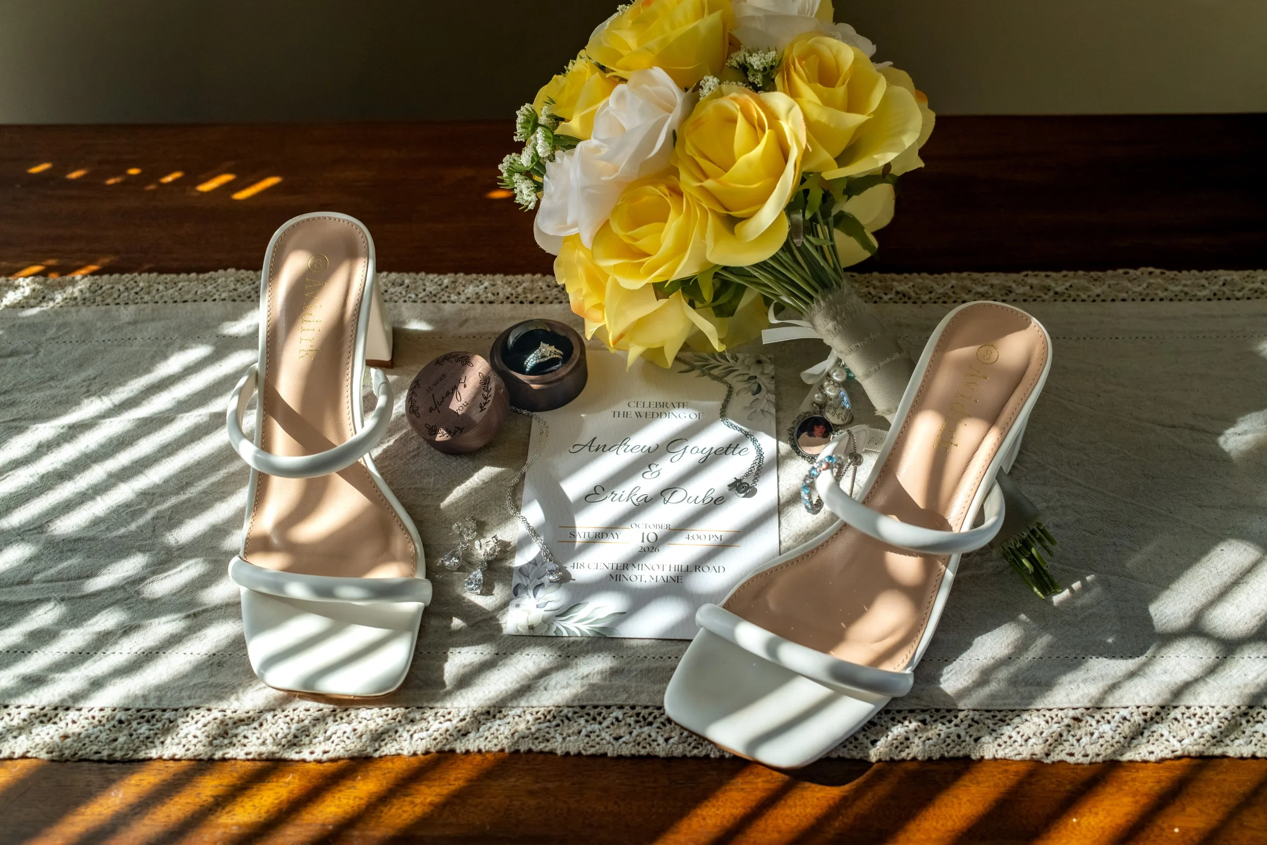 Erika's white sandals, yellow rose bouquet, and invitation suite laid out with her ring boxes on a lace runner at the Maine Wedding Barn and Event Center in Minot Maine fall wedding photography VMG Productions