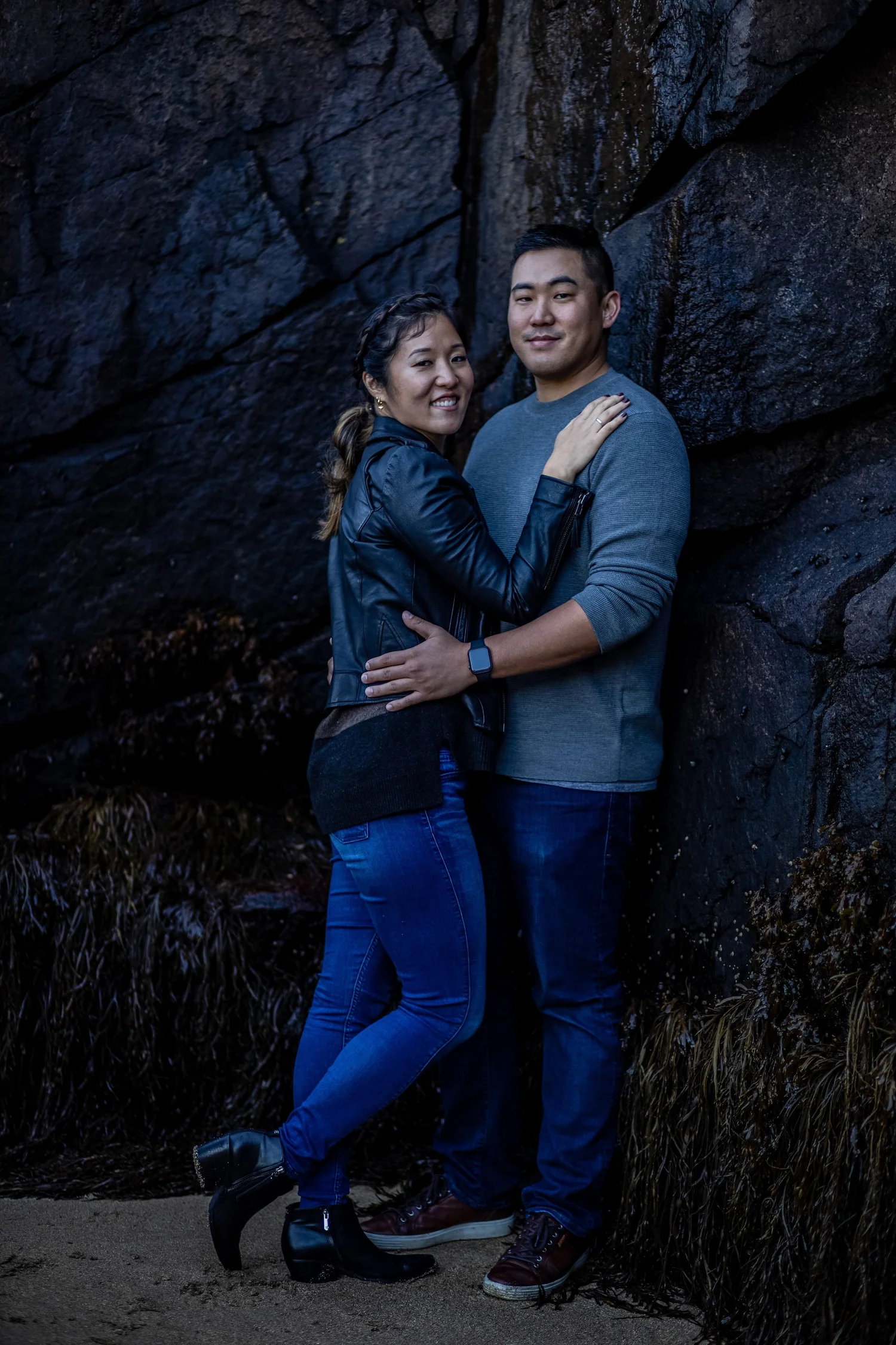 Agnes and Peter embracing against dark granite rock wall smiling at camera Acadia National Park engagement photography VMG Productions