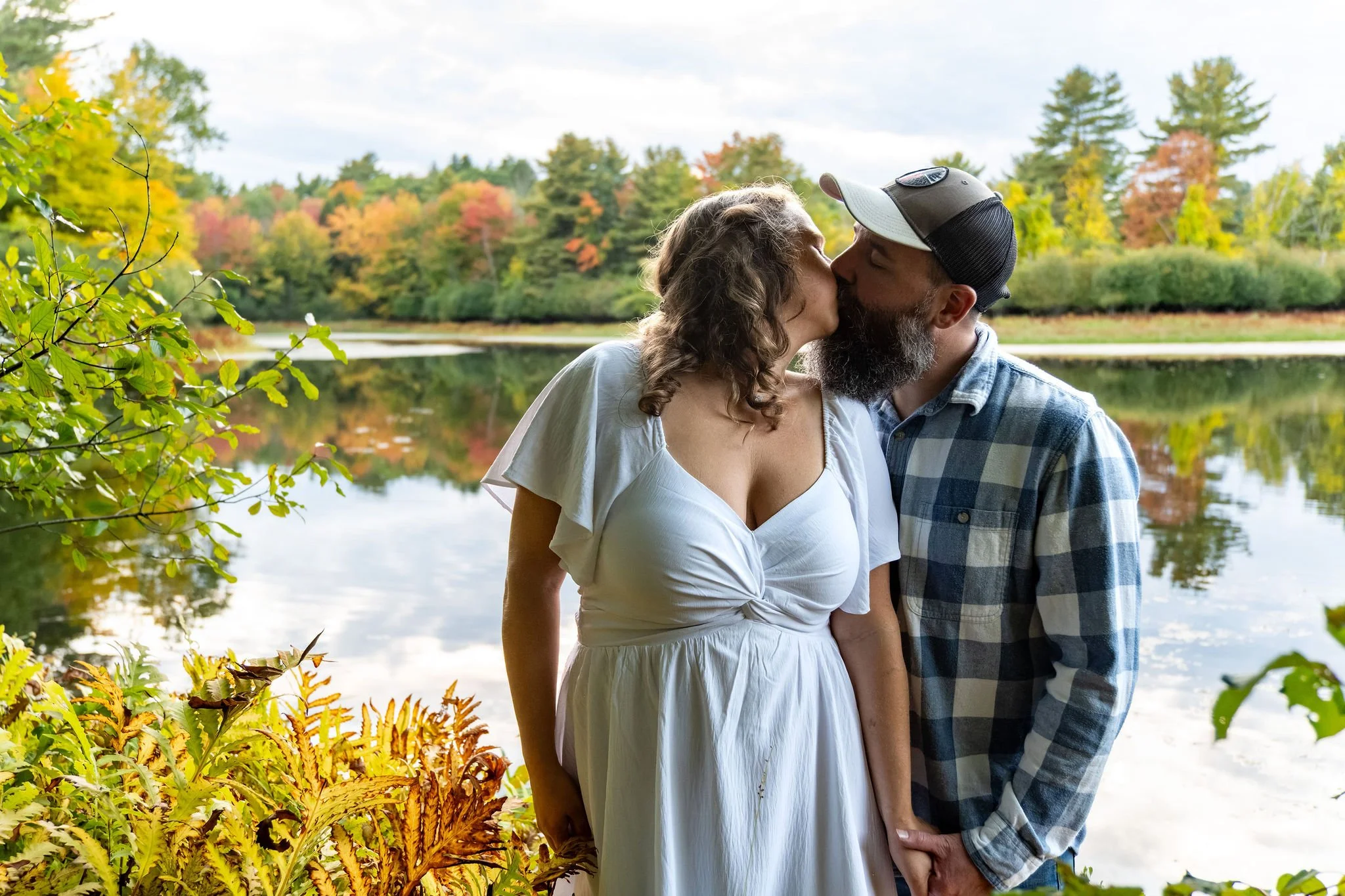Alison and Dave kissing among golden ferns with the fall shoreline reflected in the pond at Vaughan Woods in Hallowell Maine fall engagement photography VMG Productions