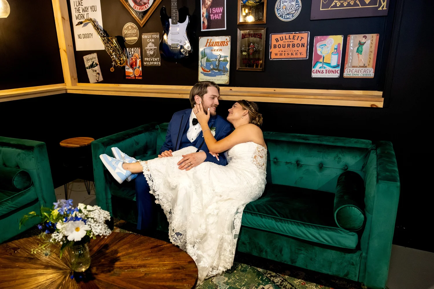 Bride and groom laughing together on velvet couch at reception showing bride's blue sneakers under wedding dress at The Barn at Hatch Point Bowdoinham Maine
