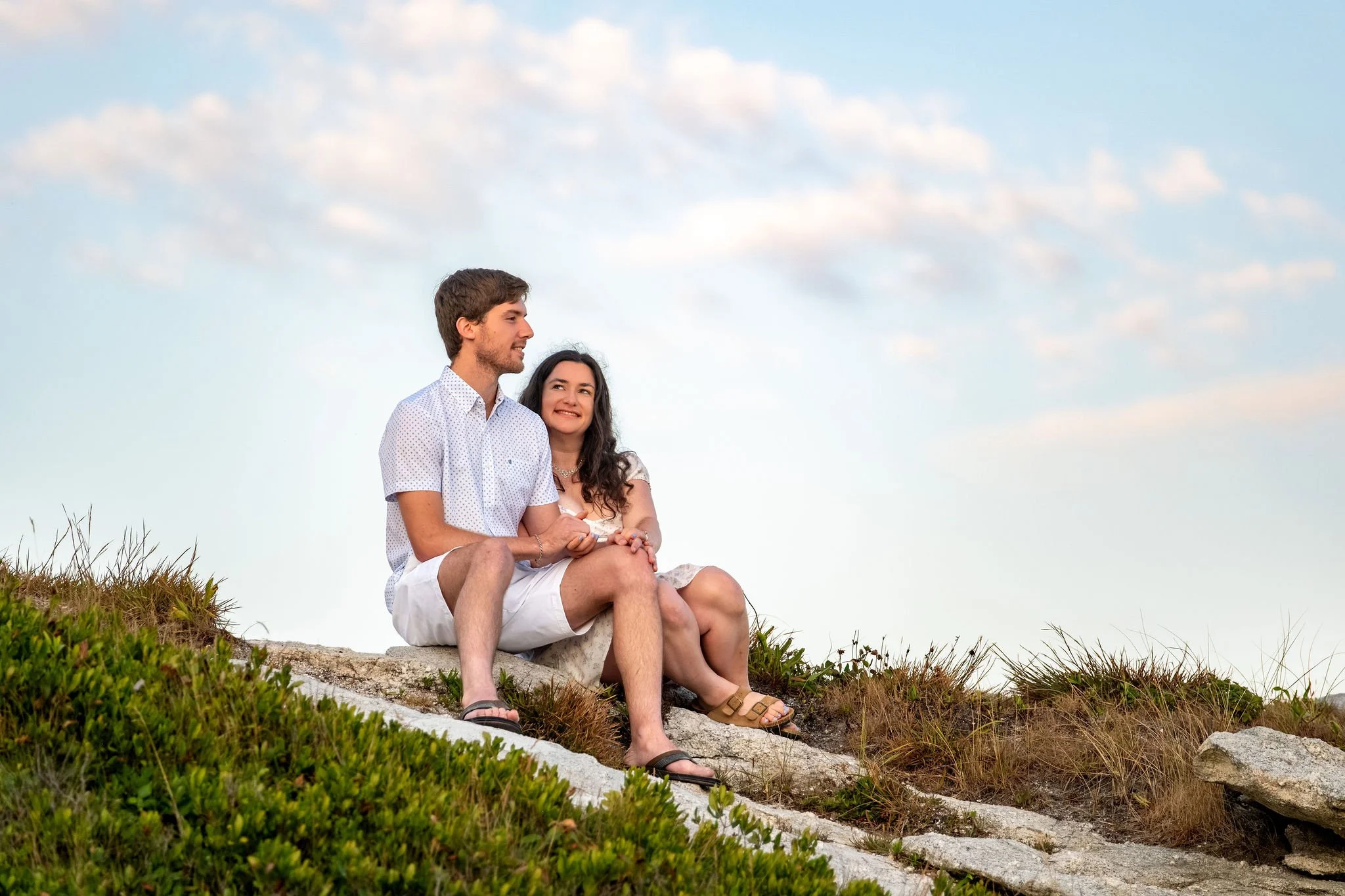 Kirstie and Lucas sitting together on the rocky headland under a sky full of clouds at Reid State Park in Georgetown Maine engagement photography VMG Productions