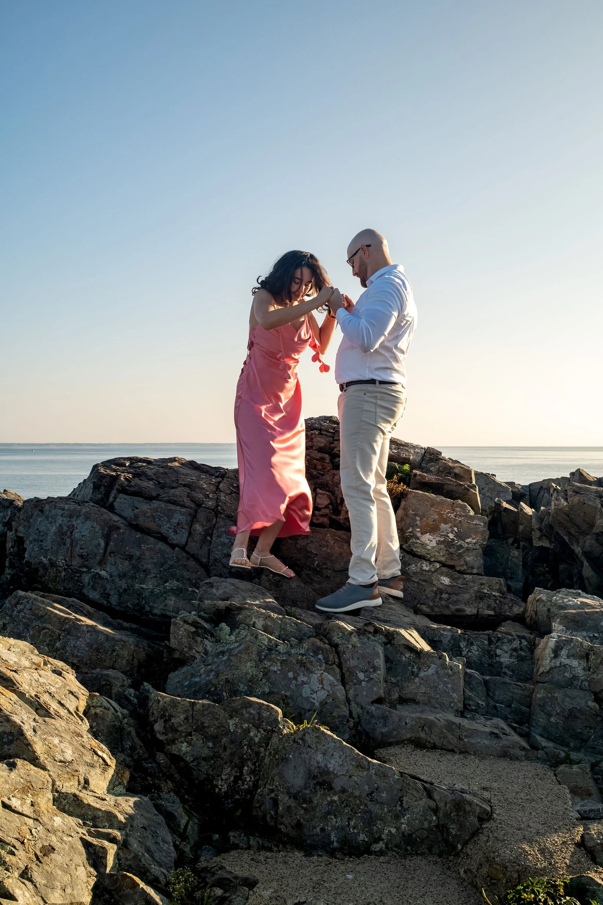 Helena stepping between the rocks with Zack's hands in hers on Marginal Way in Ogunquit Maine sunrise proposal and engagement photography VMG Productions