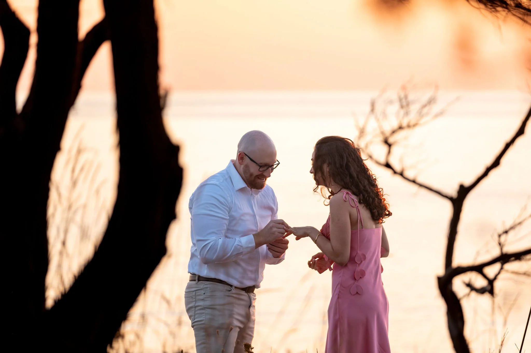 Zack placing the ring on Helena's finger beside a silhouetted tree with the sunrise glowing over the ocean behind them on Marginal Way in Ogunquit Maine sunrise proposal and engagement photography VMG Productions