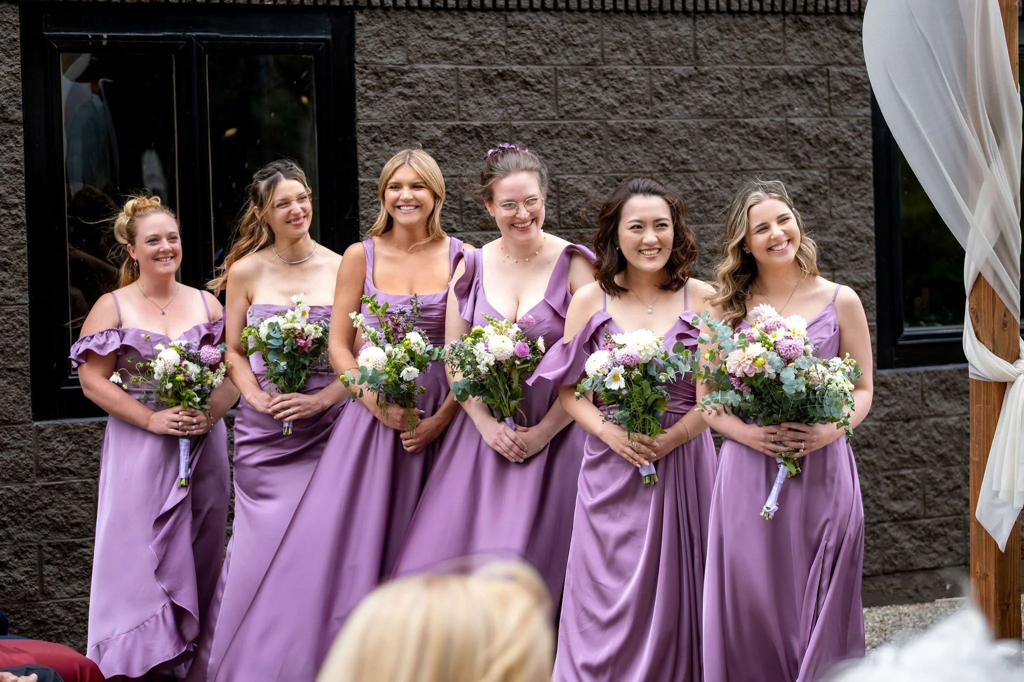 Six bridesmaids in lavender satin smiling during the ceremony against the brick wall at The Cellars at Allagash Brewing Company in Portland Maine fall wedding photography VMG Productions