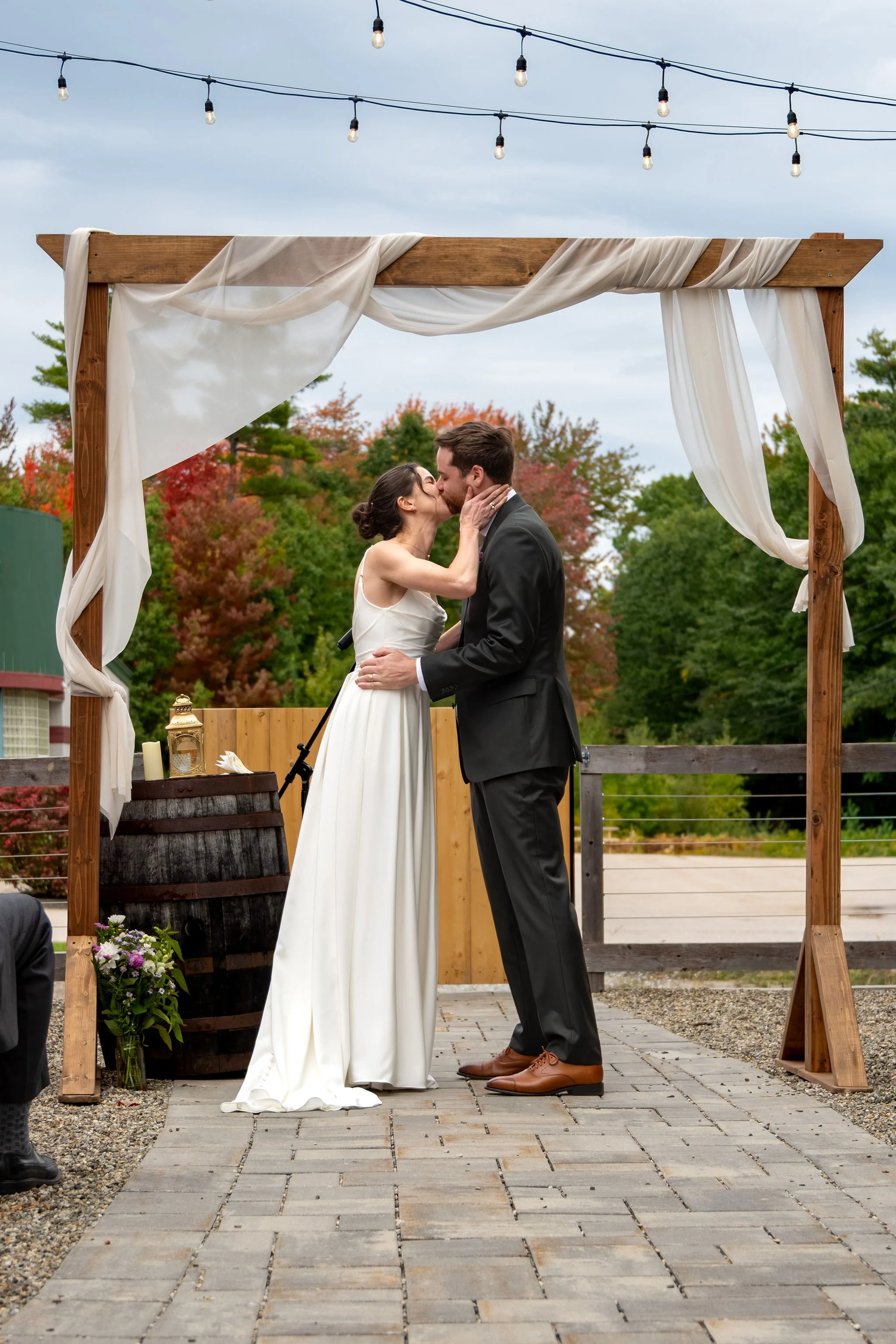 Victoria and Jack's first kiss as a married couple beneath the draped wooden arbor and string lights at The Cellars at Allagash Brewing Company in Portland Maine fall wedding photography VMG Productions