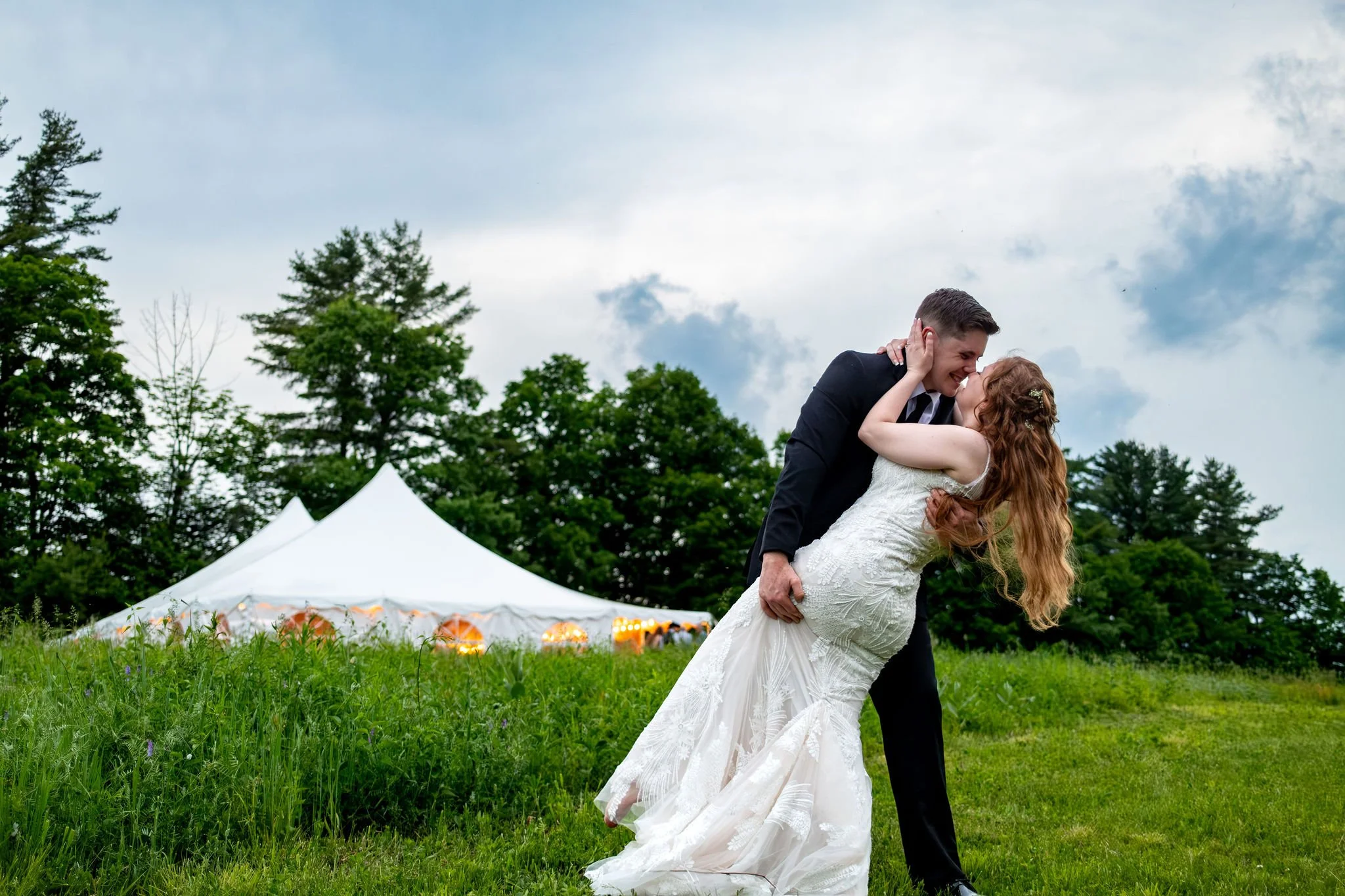 Kayla and Ethan DeAngelis dip kiss in the summer field with the lit reception tent glowing behind them at their Sanbornton New Hampshire wedding photography VMG Productions