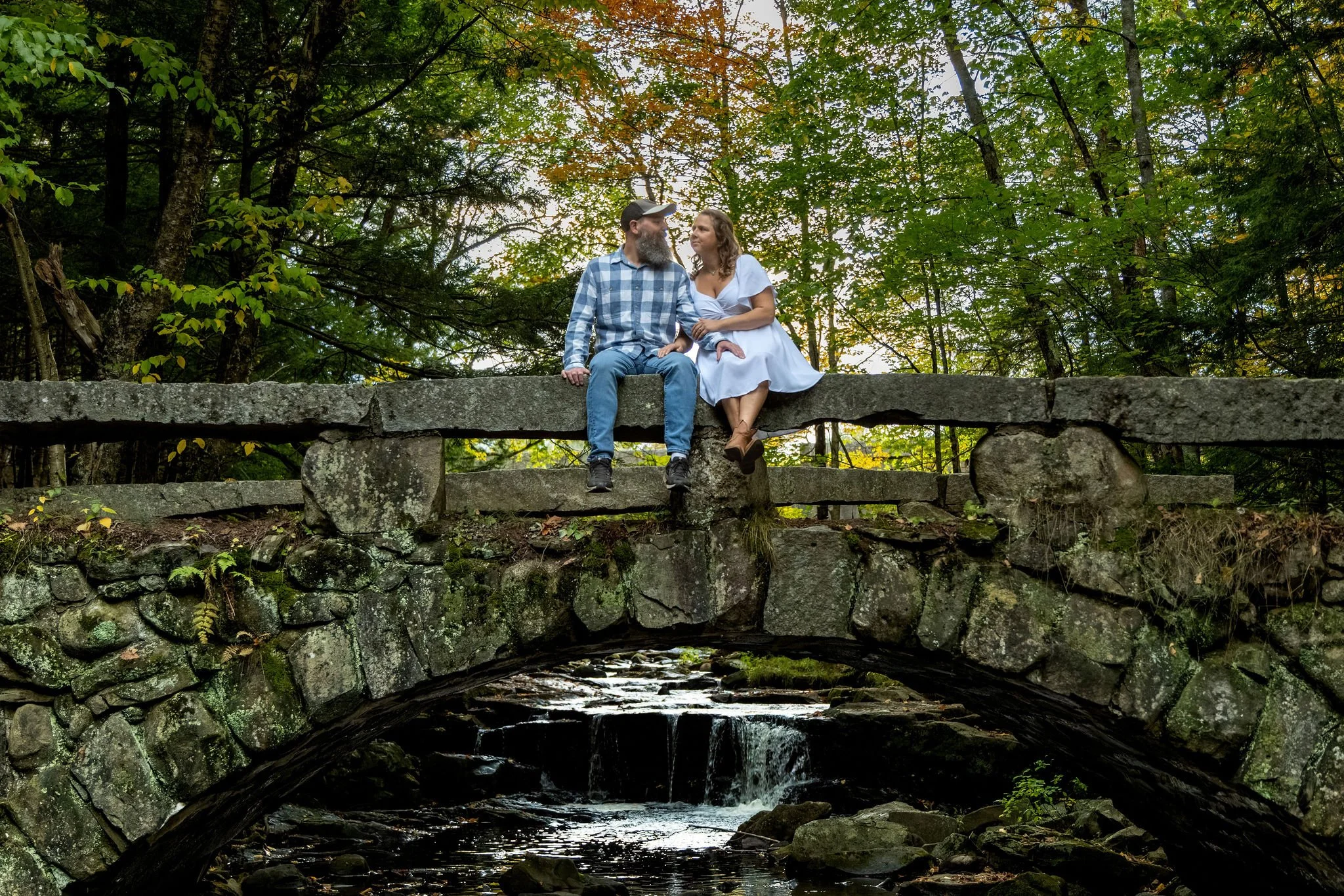 Alison and Dave sitting together on top of the granite arch bridge with the brook falling through the arch below and the maples turning overhead at Vaughan Woods in Hallowell Maine fall engagement photography VMG Productions