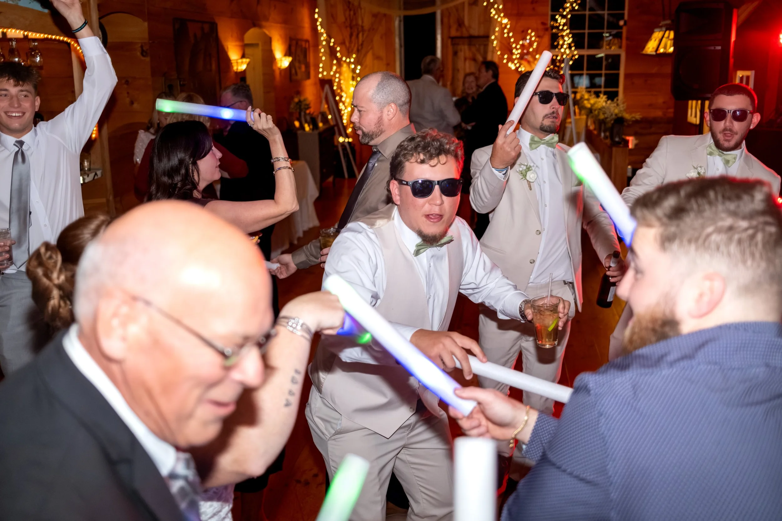Andrew in sunglasses dancing with light-up wands next to an older guest who is laughing at the Maine Wedding Barn and Event Center in Minot Maine fall wedding photography VMG Productions