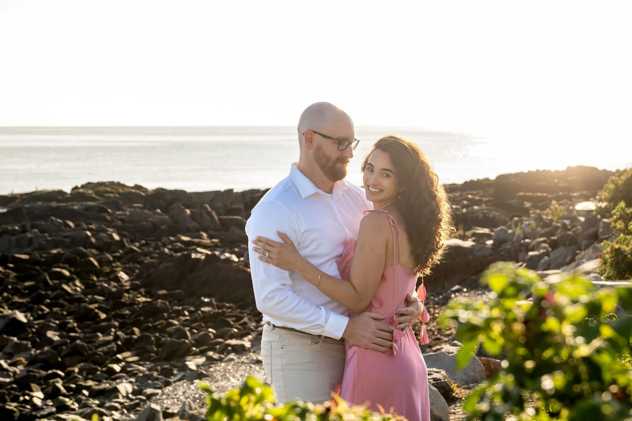 Zack and Helena smiling in a warm embrace with the sun glowing on the water behind them on Marginal Way in Ogunquit Maine sunrise proposal and engagement photography VMG Productions