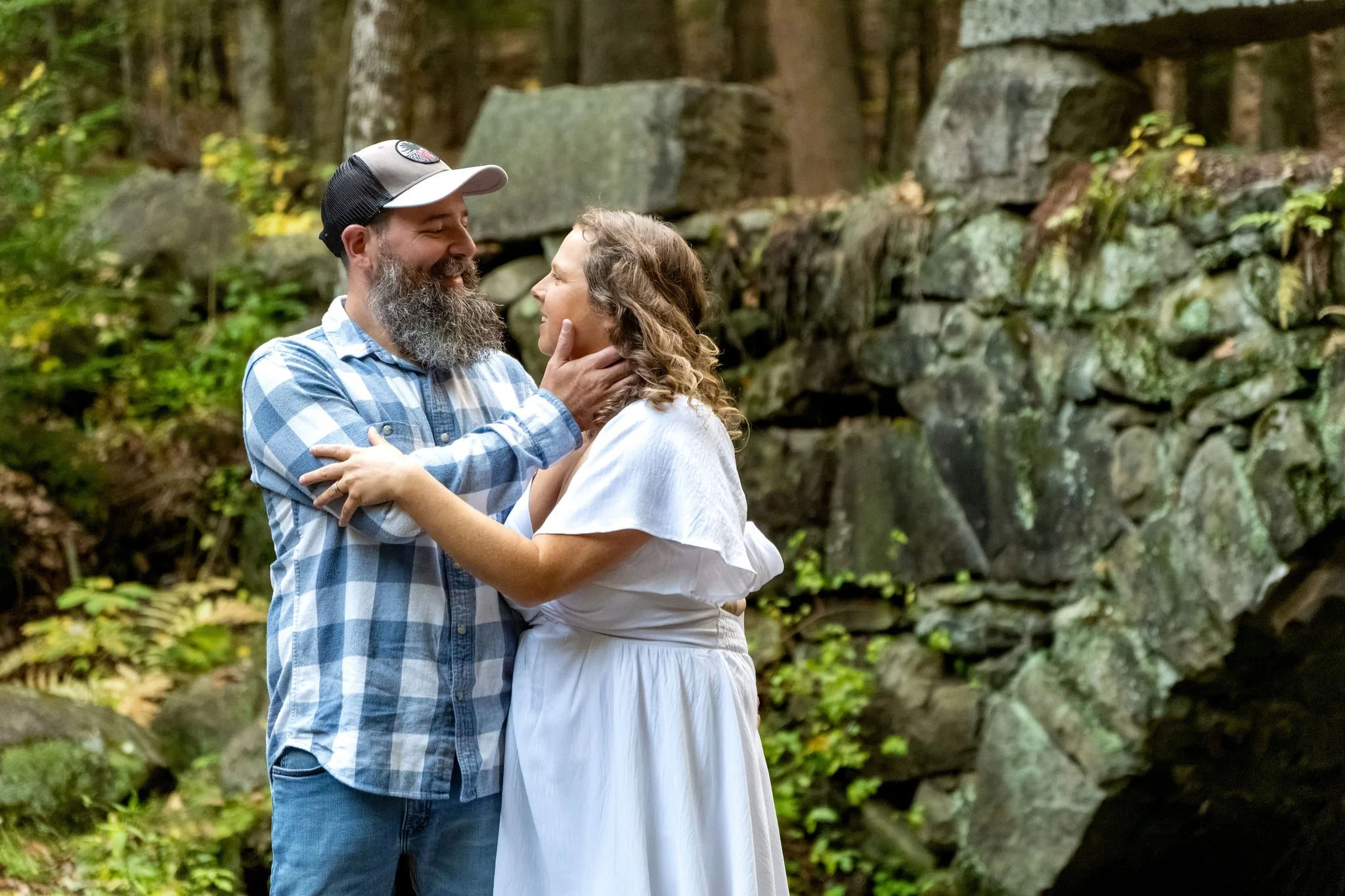 Dave holding Alison's face in his hands as they laugh together in front of the mossy stone wall at Vaughan Woods in Hallowell Maine fall engagement photography VMG Productions