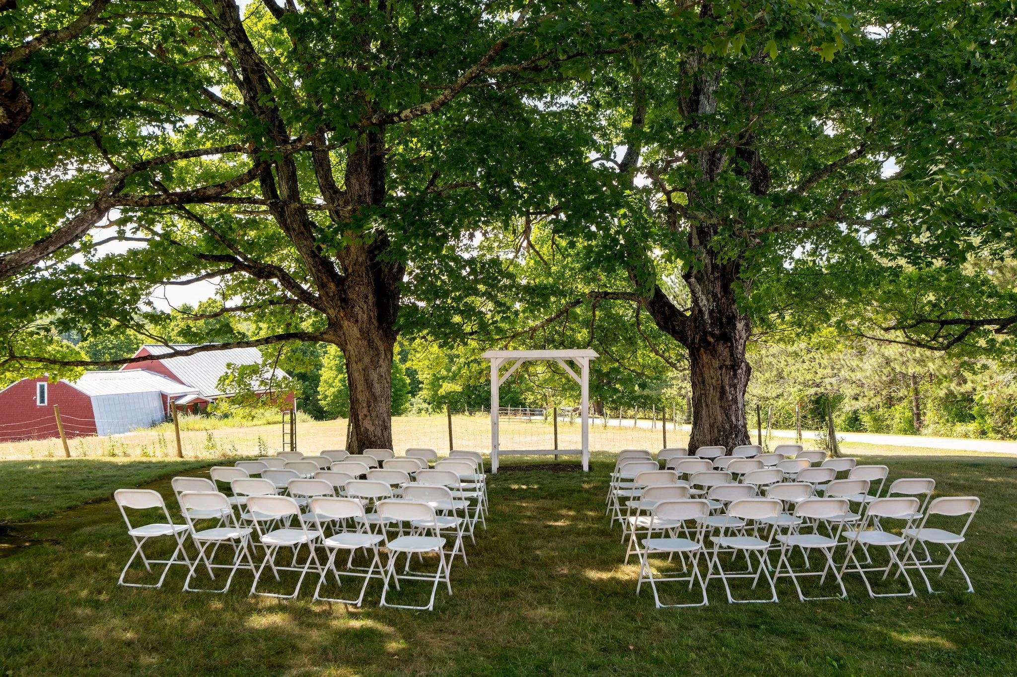 Ceremony site set with white chairs and a white arbor under two maple trees at Cassidy and Chase Begin's wedding at Maple Hill Farm Inn and Conference Center in Hallowell Maine summer wedding photography VMG Productions