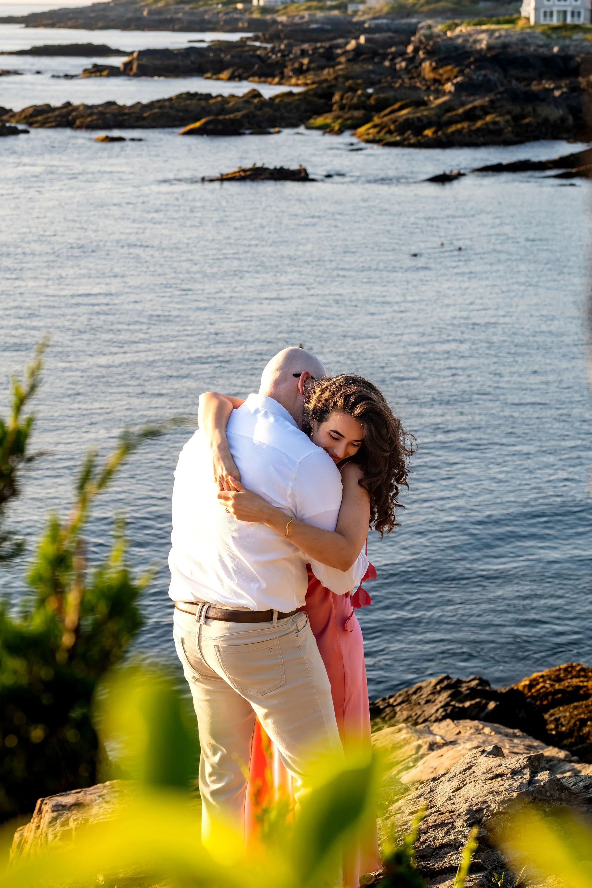 Helena resting her head on Zack's shoulder as they hug on the rocks with the coastline behind them on Marginal Way in Ogunquit Maine sunrise proposal and engagement photography VMG Productions