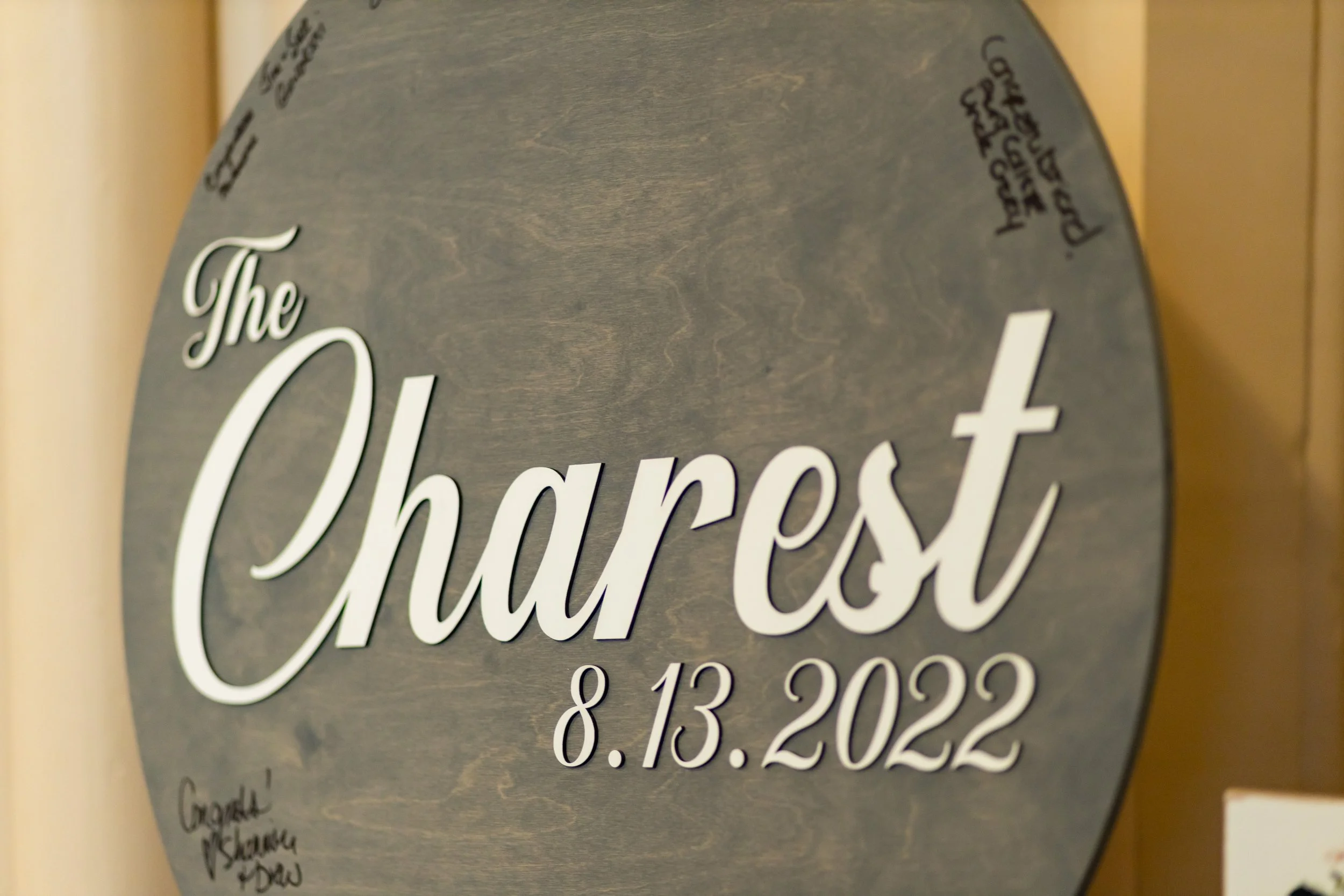 Round engraved dark wood wedding guest book sign reading The Charest 8.13.2022 with guest signatures written around the edges at Agora Grand Event Center Lewiston Maine wedding August 2022