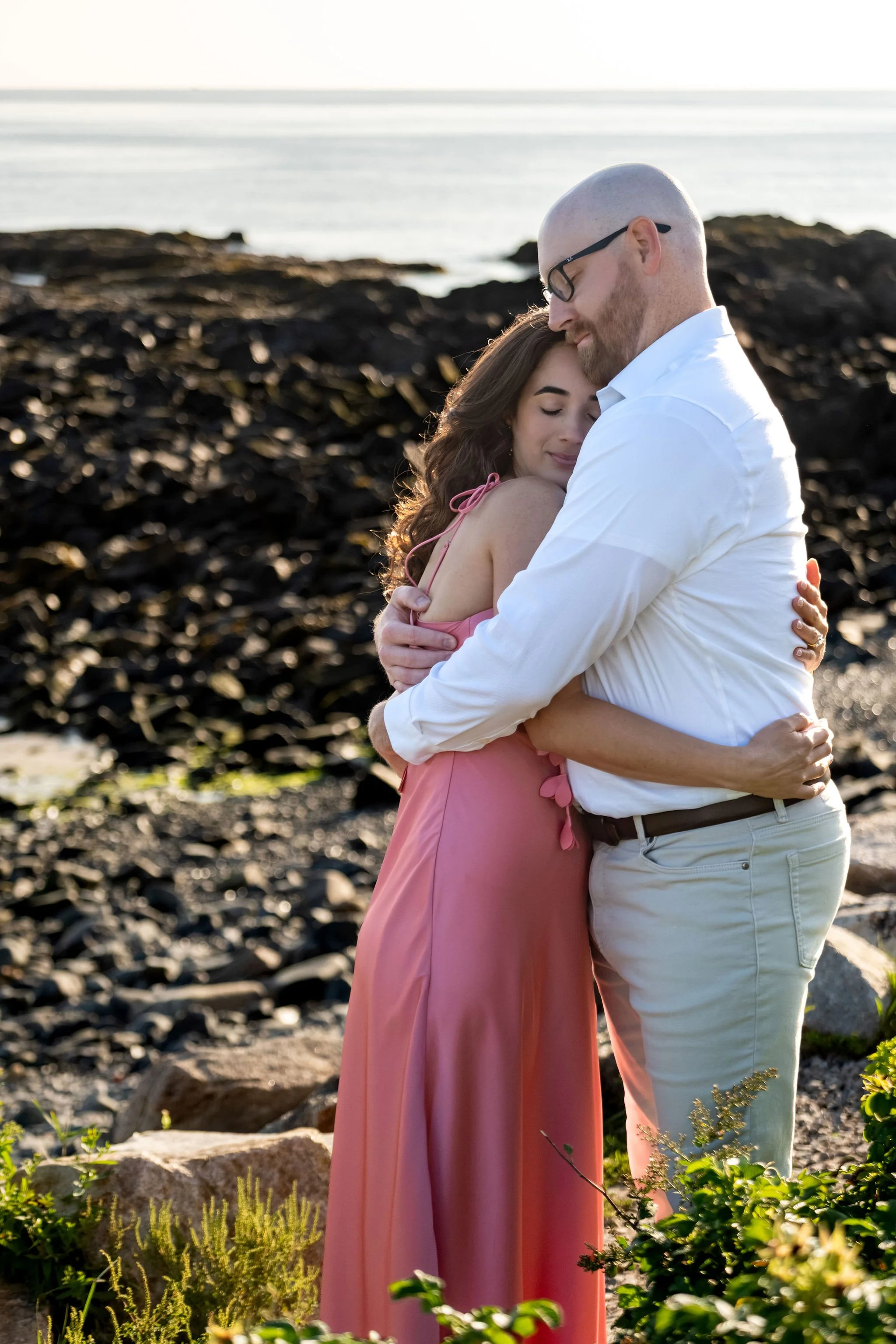 Zack kissing Helena's forehead as they hug tightly on the rocky shore on Marginal Way in Ogunquit Maine sunrise proposal and engagement photography VMG Productions
