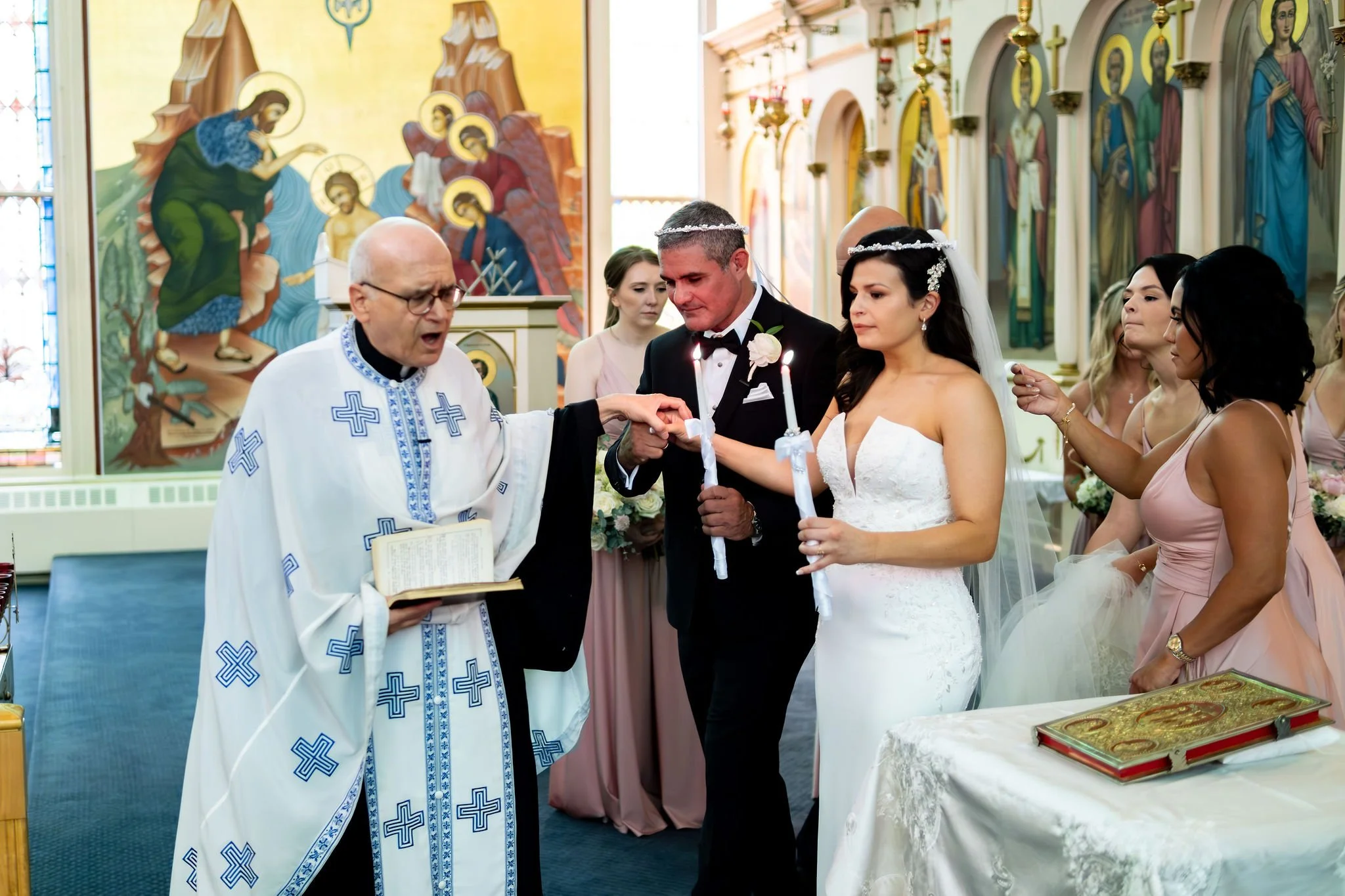 The priest joining Nicole and Alexey's hands as they hold their candles at Holy Trinity Greek Orthodox Church Portland Maine summer wedding photography VMG Productions