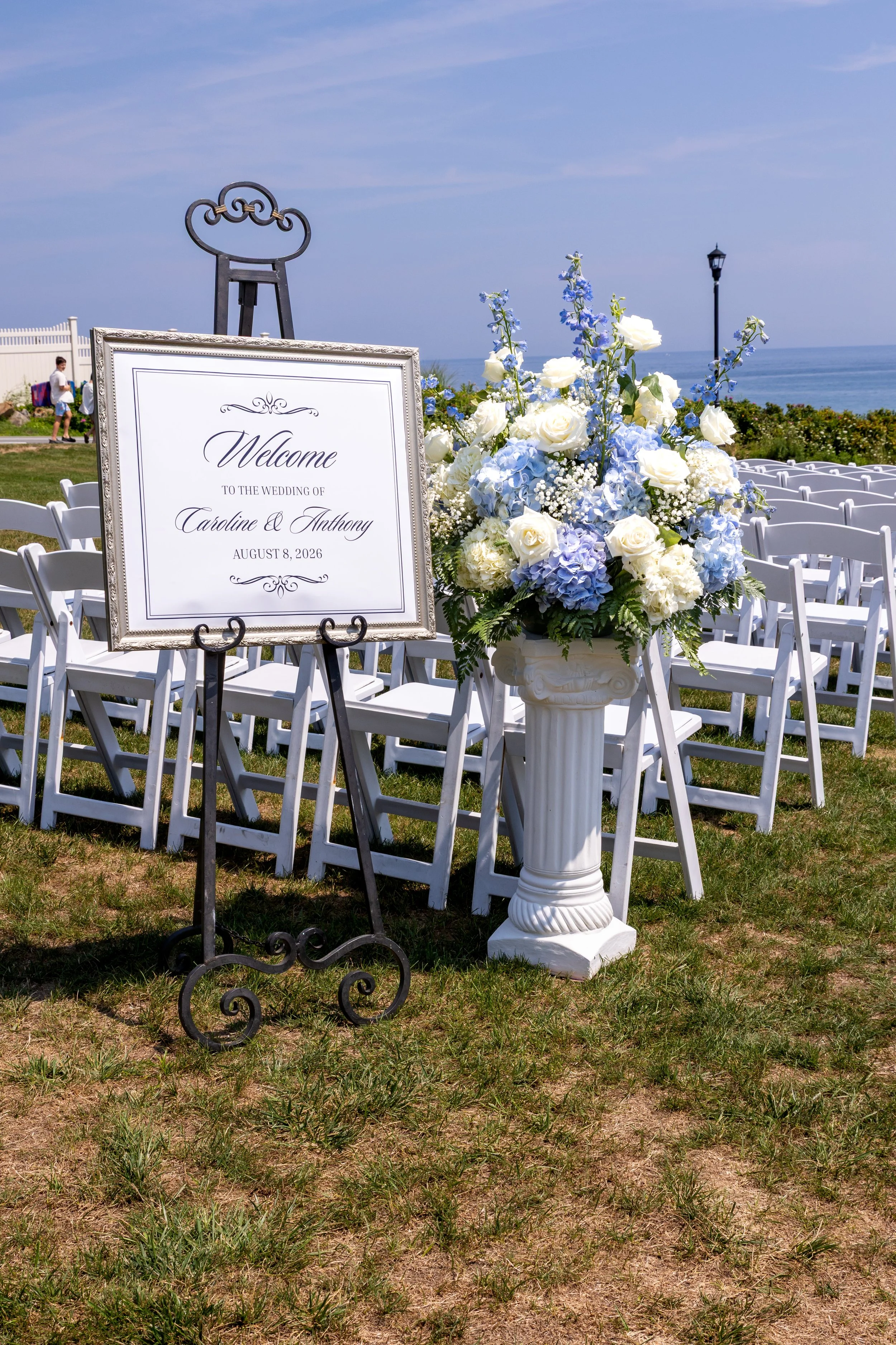 Welcome sign for the wedding of Caroline and Anthony beside a pedestal of blue hydrangeas and white roses with the ocean behind at Union Bluff Hotel in York Maine wedding photography VMG Productions