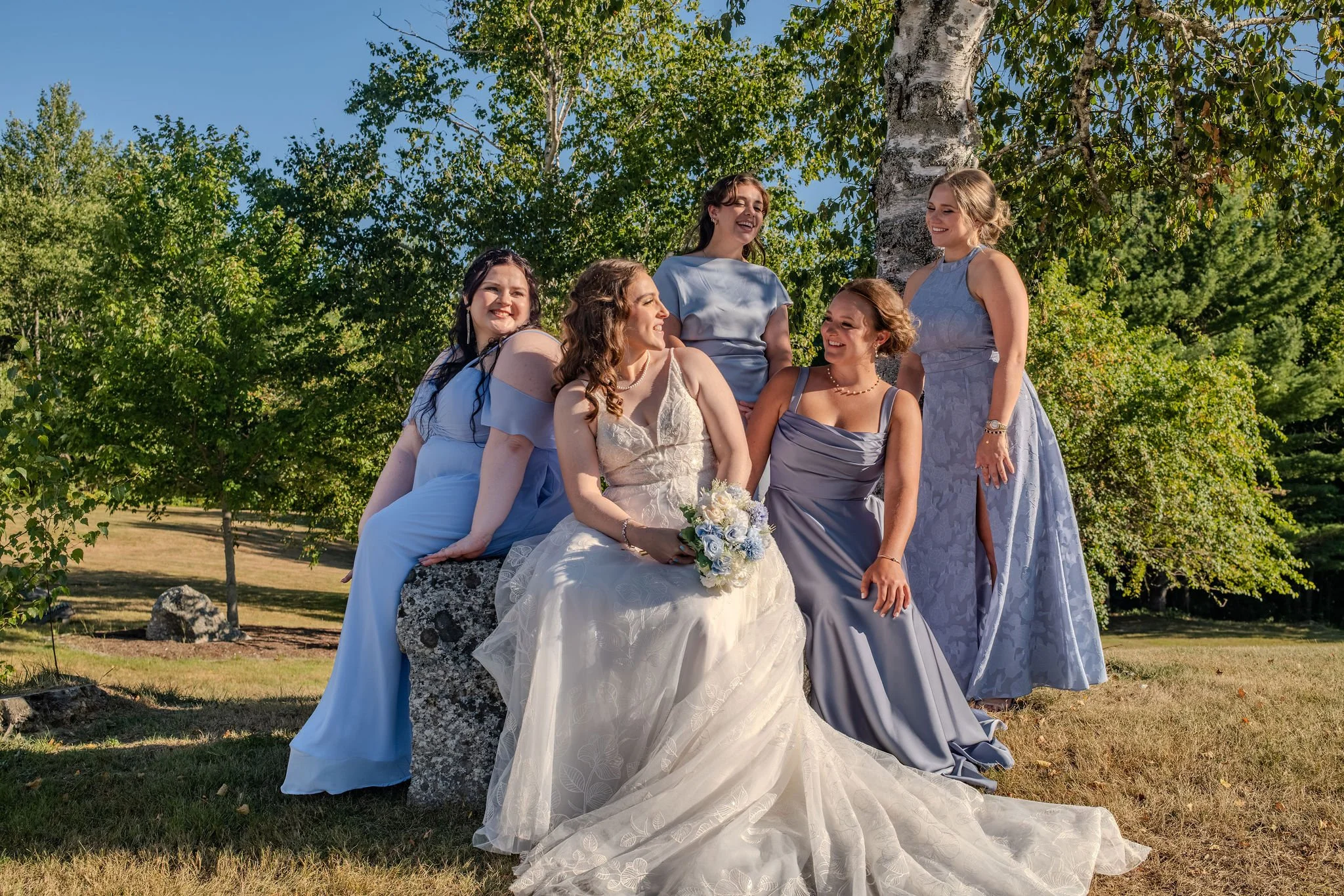 Hollie with her bridesmaids in shades of blue seated on a granite boulder under the birches at The Lucerne Inn in Dedham Maine summer wedding photography VMG Productions