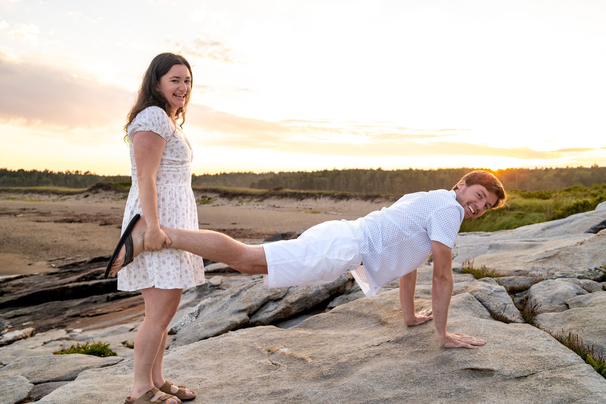 Lucas doing a plank on the rocks while Kirstie looks on unimpressed at Reid State Park in Georgetown Maine engagement photography VMG Productions