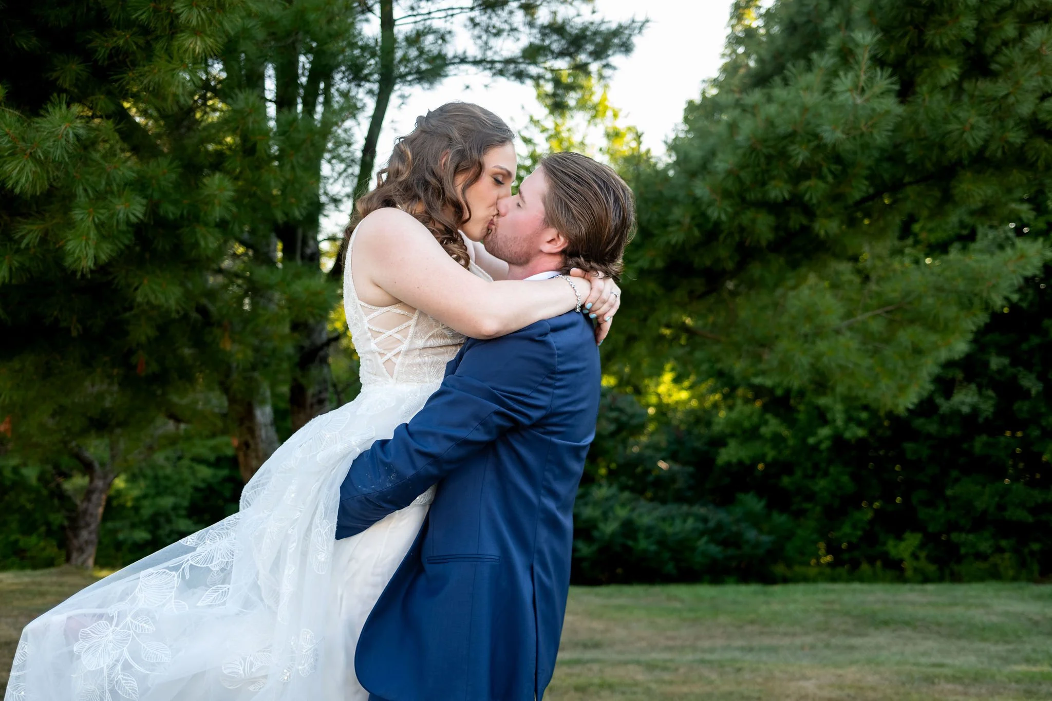Zachary lifting Hollie off the ground for a kiss in front of the pines with her dress swept up at The Lucerne Inn in Dedham Maine summer wedding photography VMG Productions