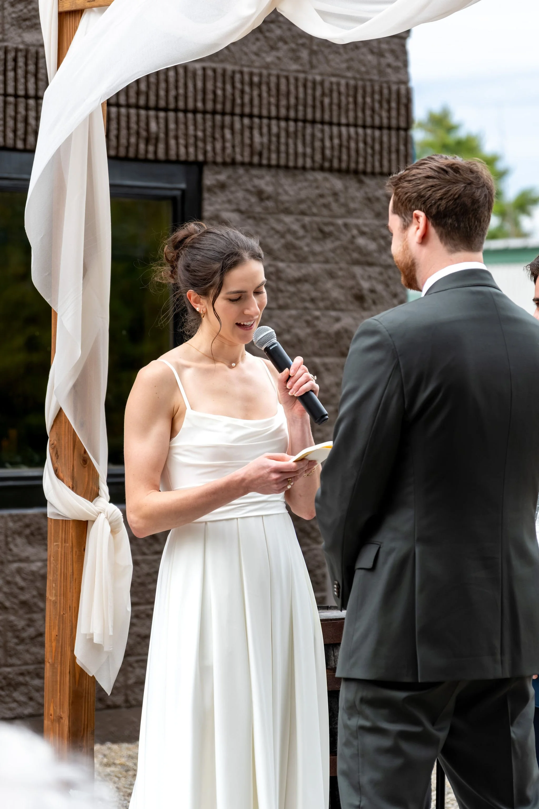 Victoria reading her vows into a microphone as Jack listens at The Cellars at Allagash Brewing Company in Portland Maine fall wedding photography VMG Productions