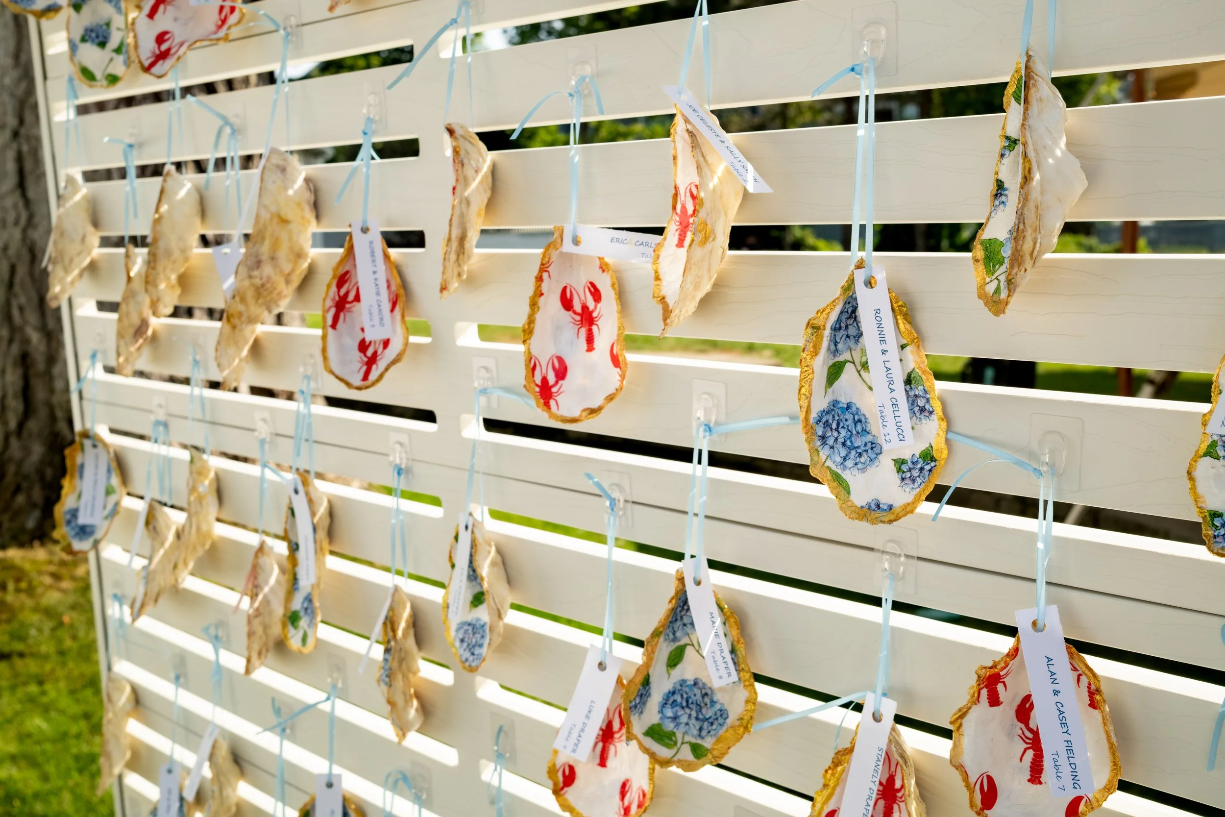 Hand painted oyster shell escort cards with lobster and blue hydrangea patterns hung with light blue ribbon at Ashley and Marc Draper's reception at The Breakwater Inn in Kennebunkport Maine summer wedding photography VMG Productions