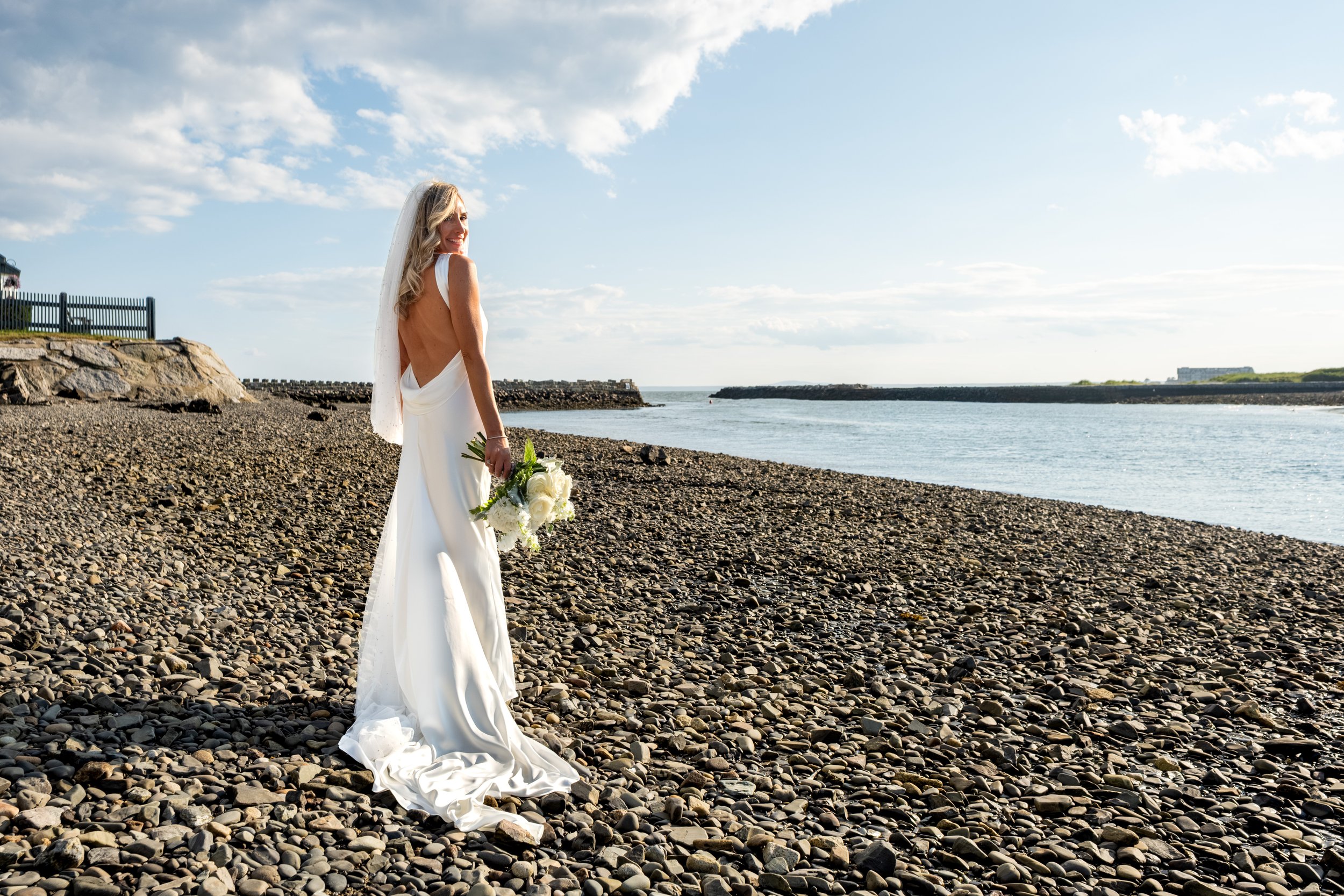 Ashley Draper glancing back over her shoulder with her pearl veil on the pebble beach at The Breakwater Inn in Kennebunkport Maine summer wedding photography VMG Productions