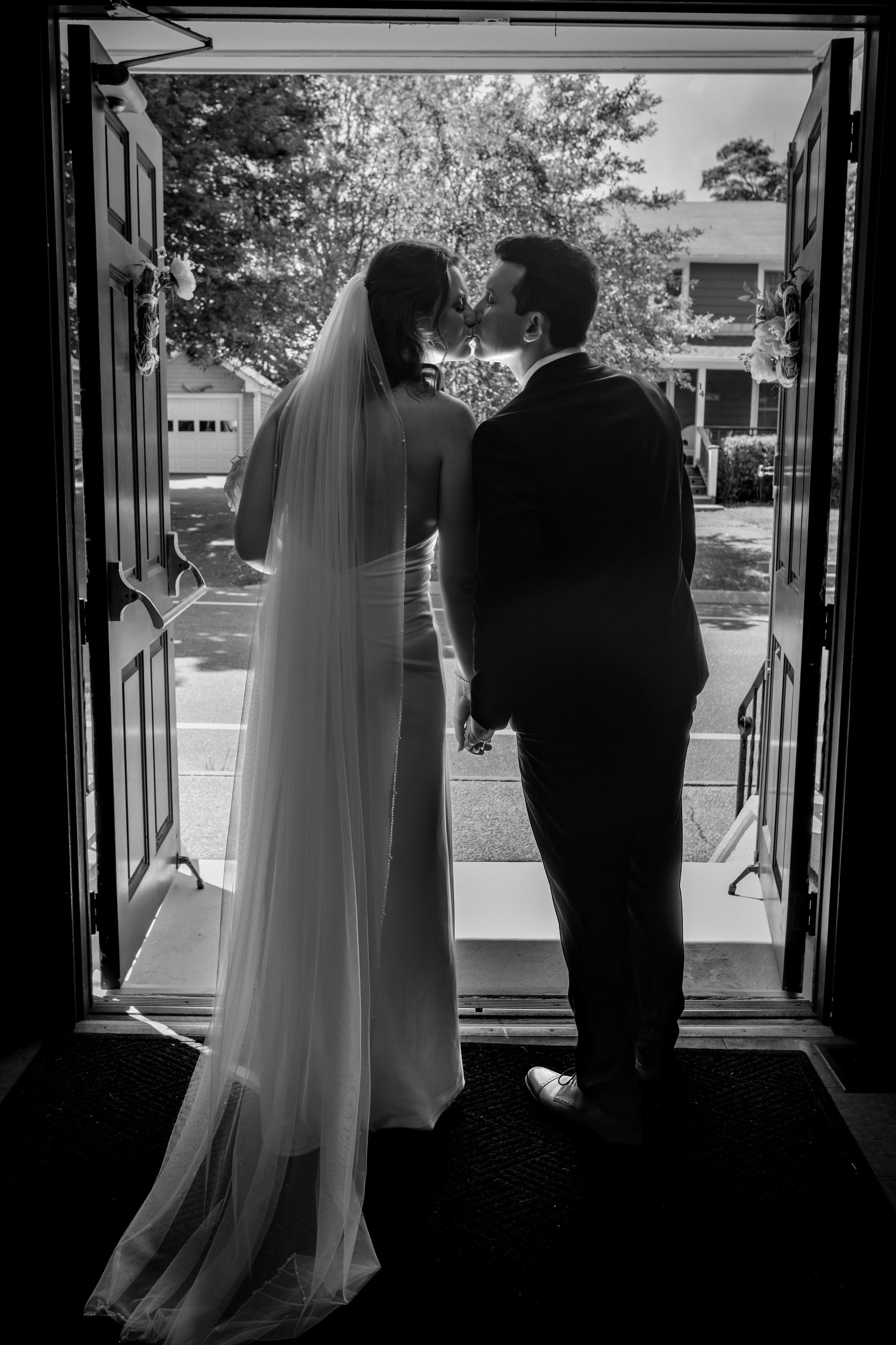 Caroline and Anthony Pirolli silhouetted in the church doorway kissing with the veil catching the light in black and white at Star of the Sea in York Maine wedding photography VMG Productions