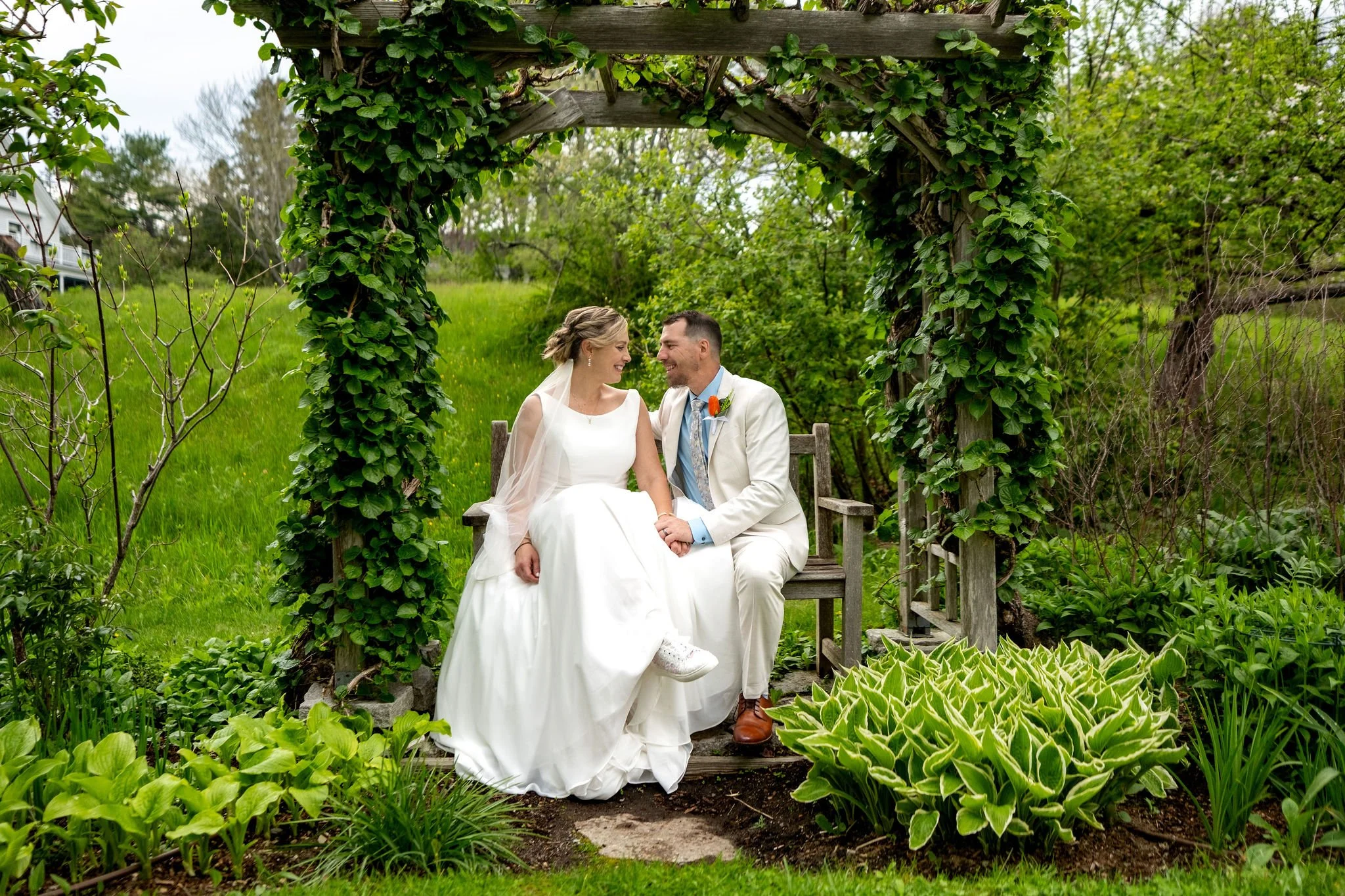 Alli and Adam Baudoin sitting together on the bench beneath the green garden arbor in Somesville on Mount Desert Island Maine spring wedding photography VMG Productions