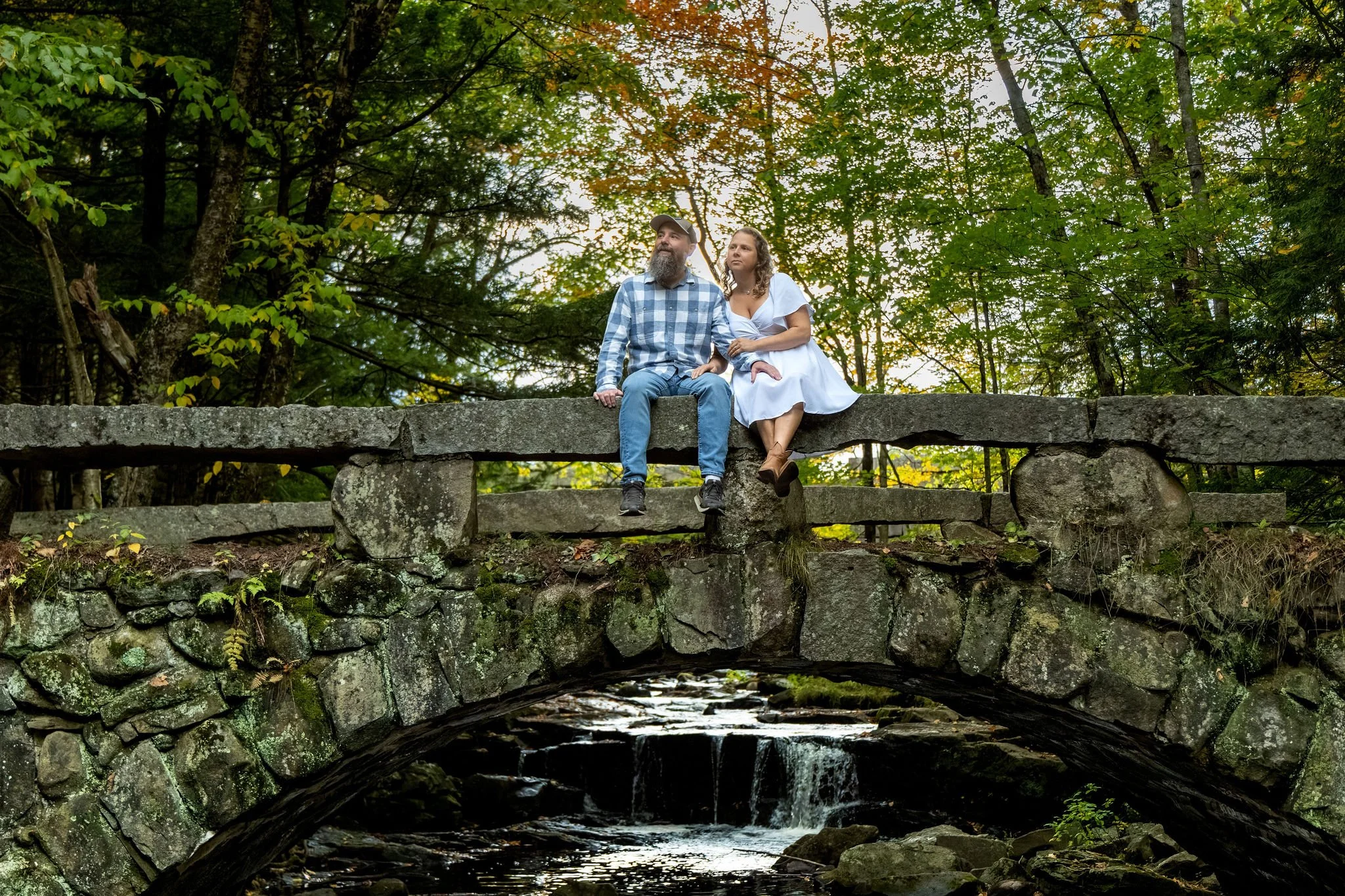Alison and Dave sitting close on the stone bridge with the arch and the falls beneath them at Vaughan Woods in Hallowell Maine fall engagement photography VMG Productions