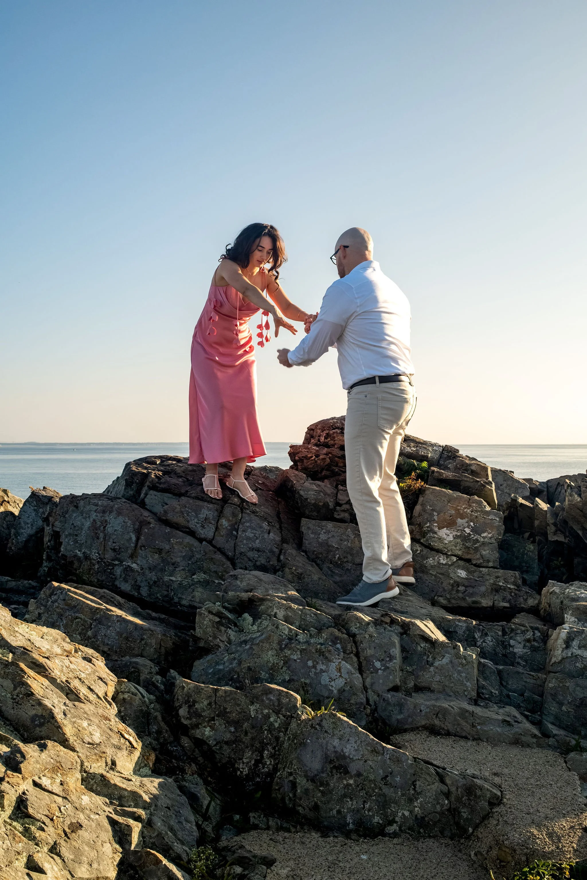Zack and Helena holding hands and laughing on the rocks with the ocean horizon behind them on Marginal Way in Ogunquit Maine sunrise proposal and engagement photography VMG Productions