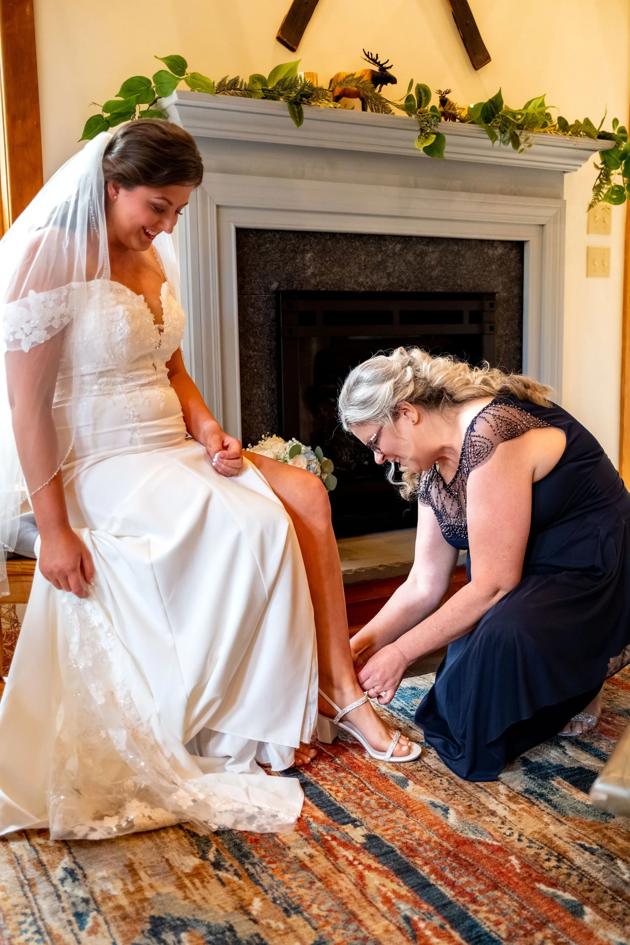 Alyssa and Colby the bride's mom fastening her shoes by the fireplace before the ceremony in Bethel Maine summer wedding photography VMG Productions