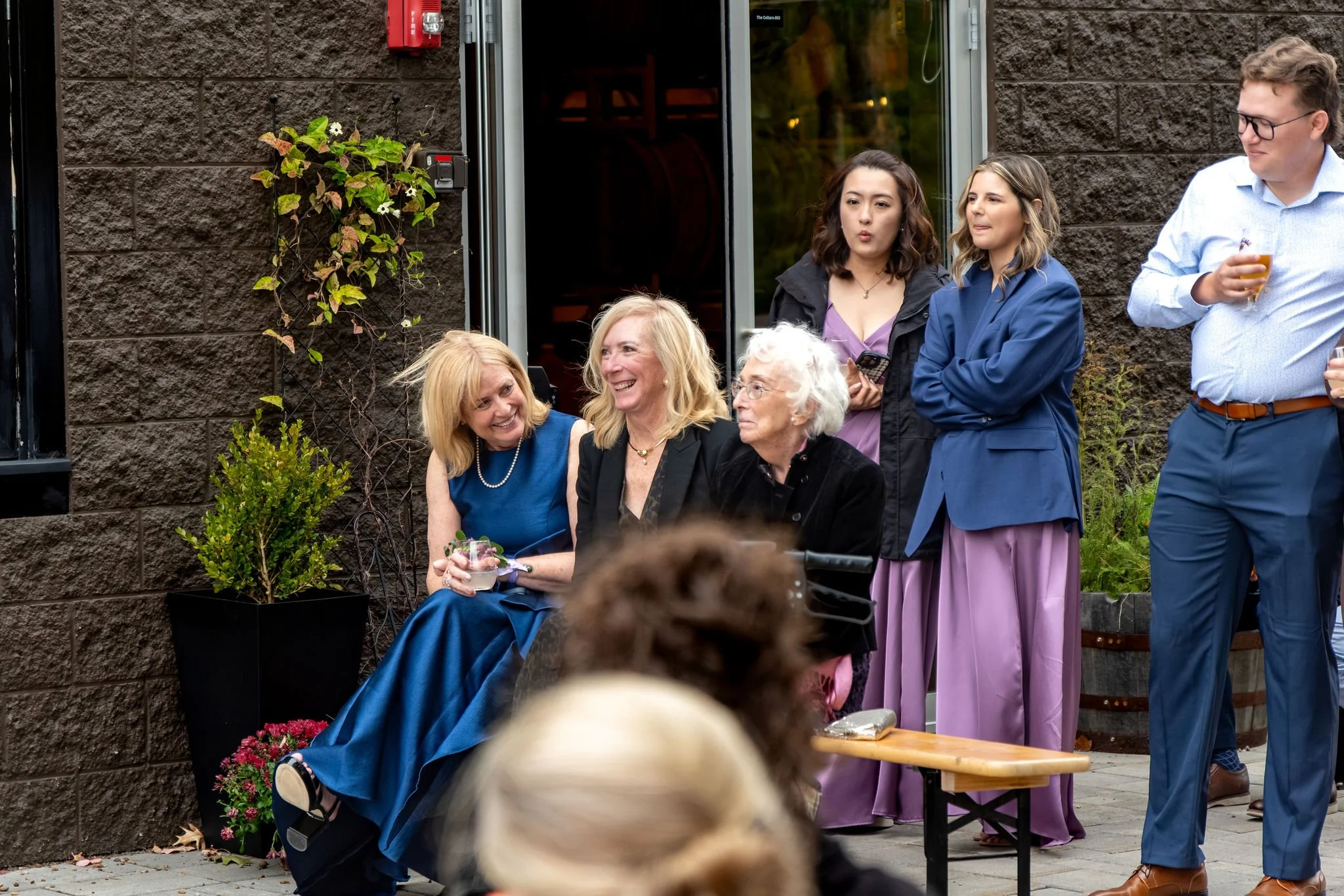 Family members laughing together on a bench by the brewery doors during the toasts at The Cellars at Allagash Brewing Company in Portland Maine fall wedding photography VMG Productions