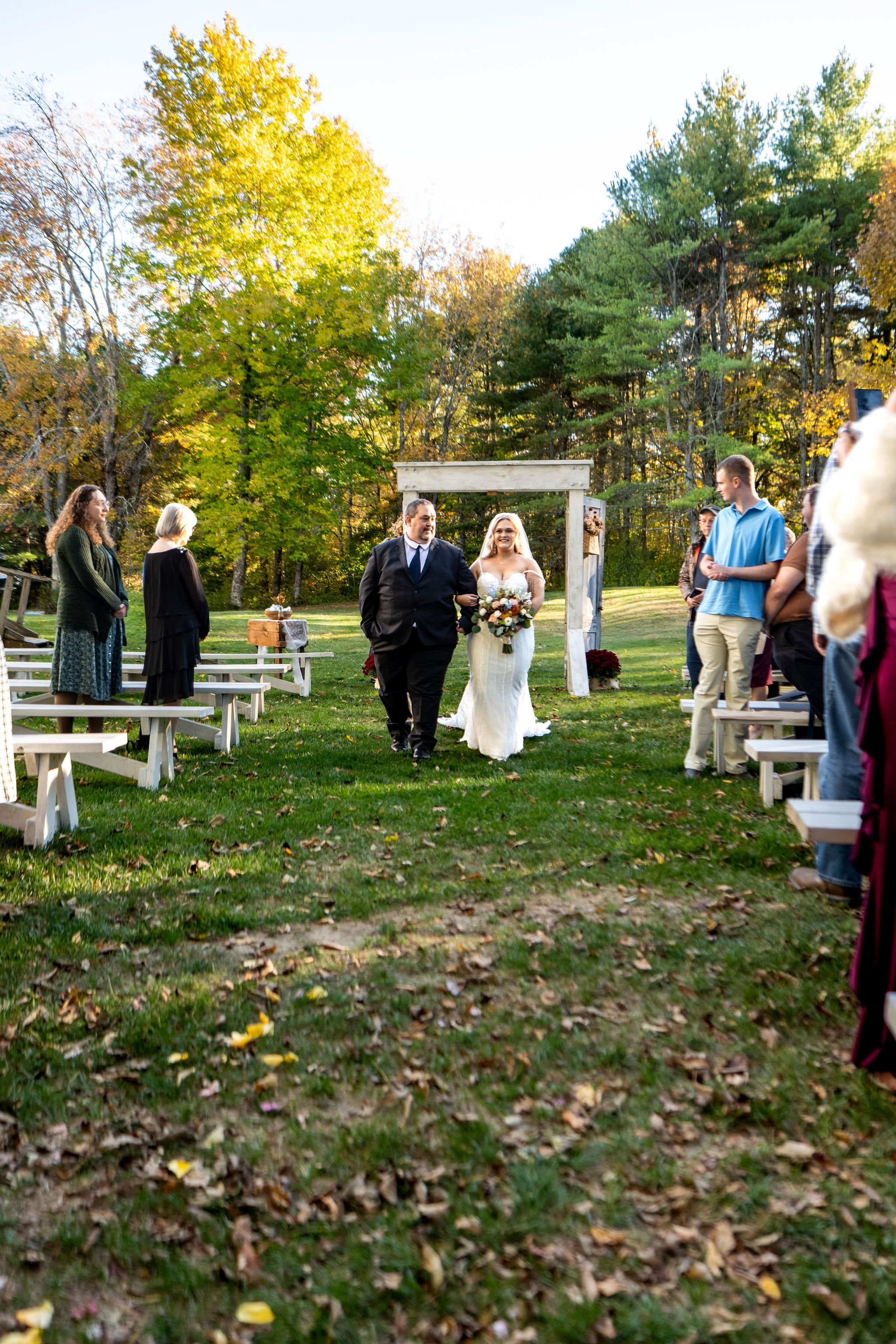 Ellie walking down the long grass aisle with her dad toward the pergola as guests stand on both sides at Boothbay Playhouse in Boothbay Maine fall wedding photography VMG Productions