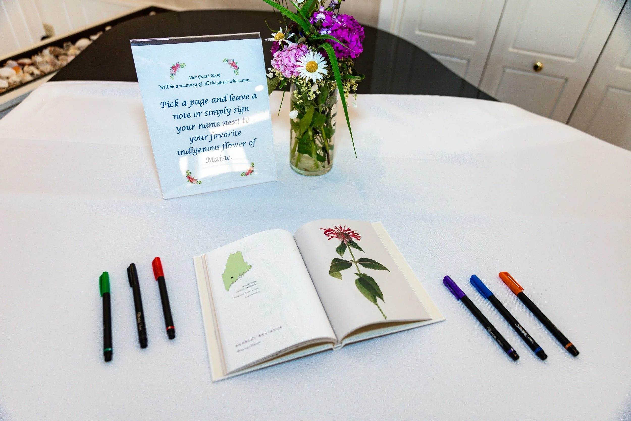Wedding guest book table at Sebasco Resort Phippsburg Maine showing Maine botanical field guide open to scarlet bee-balm illustration with prompt sign reading pick a page and sign your name next to your favorite indigenous flower of Maine wildflower bouquet and colorful fine tip pens