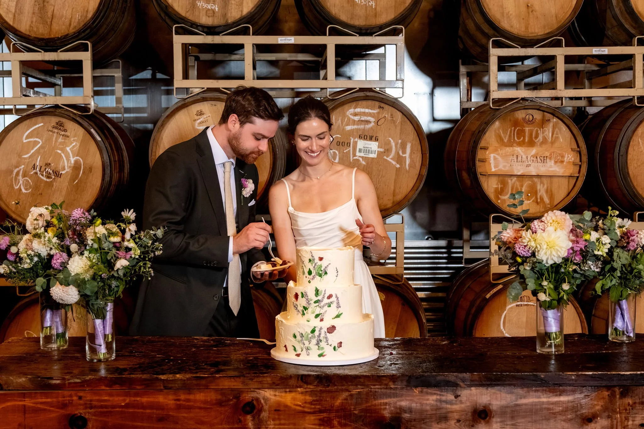 Victoria and Jack cutting their painted wedding cake at the wooden bar with wildflowers on either side at The Cellars at Allagash Brewing Company in Portland Maine fall wedding photography VMG Productions