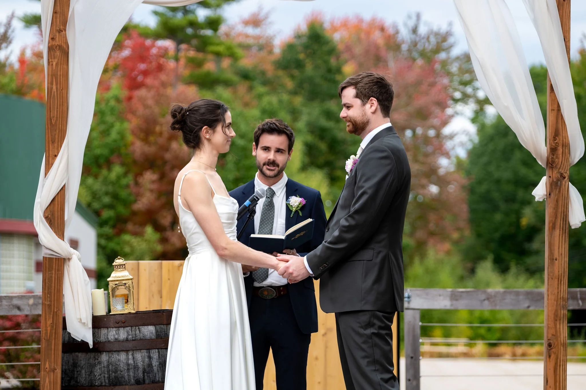 Victoria and Jack holding hands under the arbor as one of the groomsmen officiates at The Cellars at Allagash Brewing Company in Portland Maine fall wedding photography VMG Productions