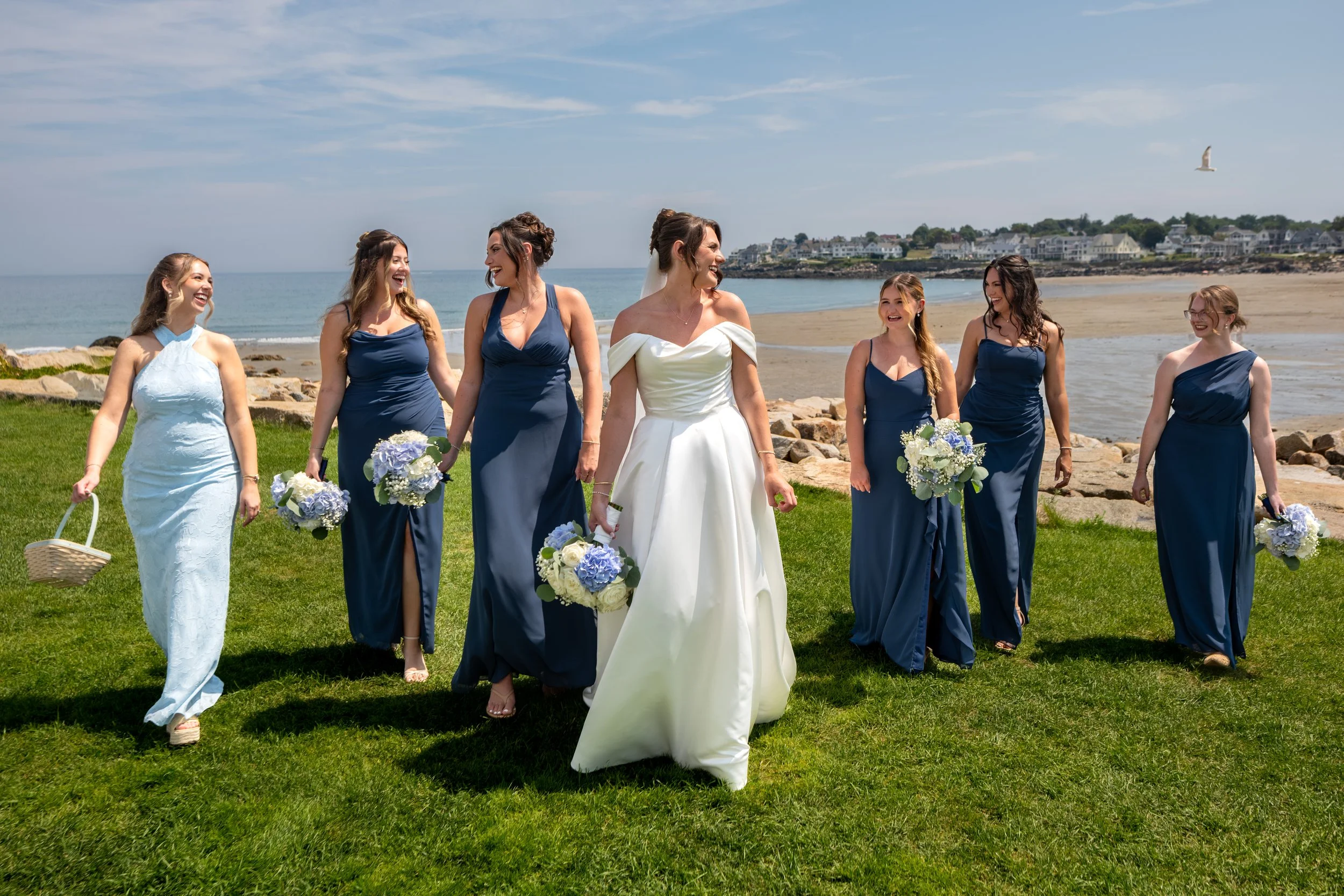 Caroline Pirolli and her bridesmaids in navy walking on the oceanfront lawn with hydrangea bouquets and the beach behind them at Union Bluff Hotel in York Maine wedding photography VMG Productions