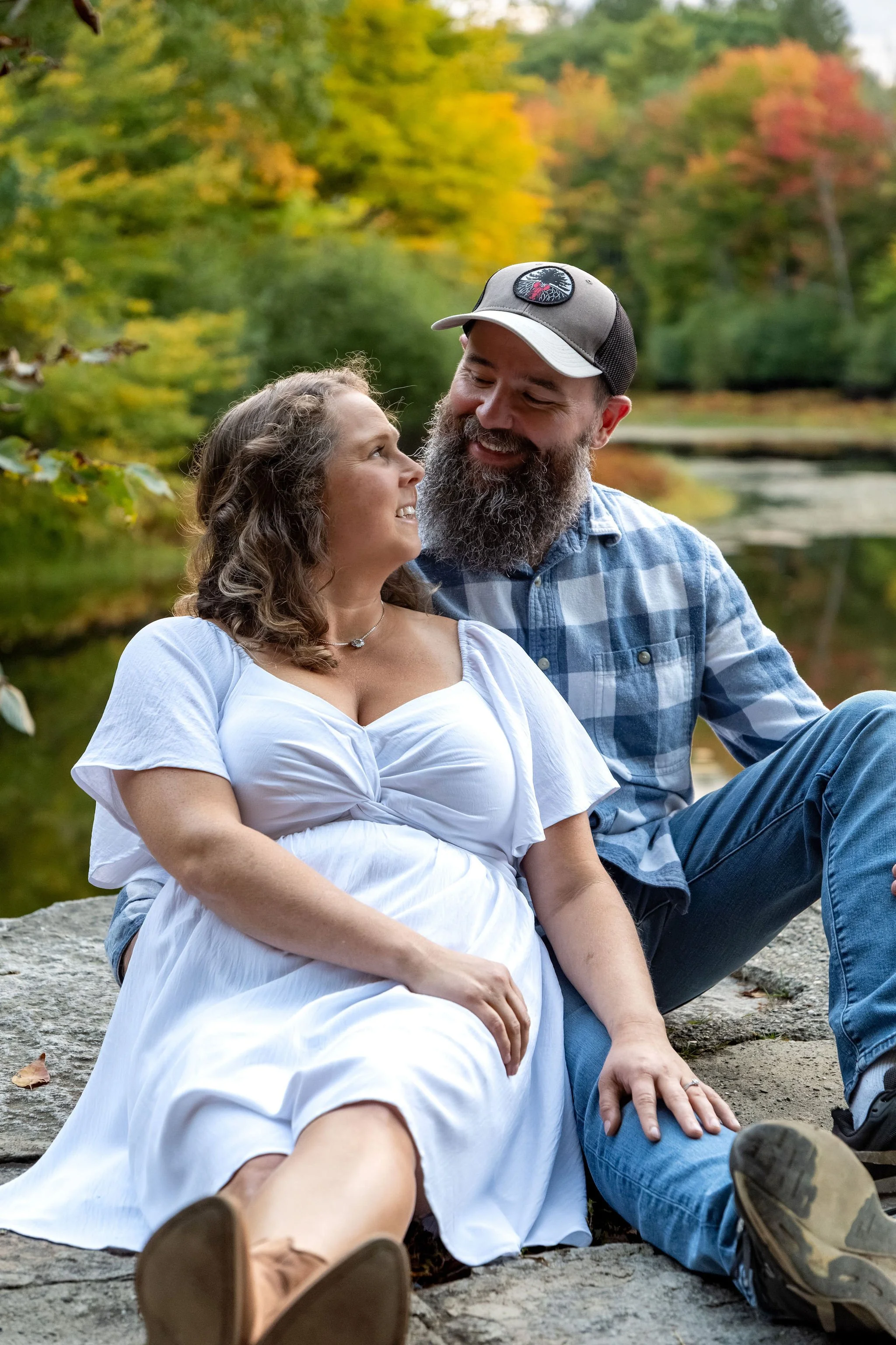 Alison and Dave sitting together on the granite at the pond's edge looking at each other with red and gold maples behind them at Vaughan Woods in Hallowell Maine fall engagement photography VMG Productions