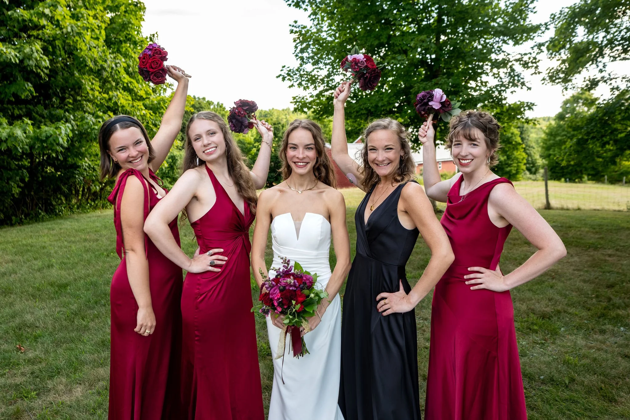 Cassidy Begin with her bridesmaids in burgundy and black raising their bouquets on the lawn at Maple Hill Farm Inn and Conference Center in Hallowell Maine summer wedding photography VMG Productions