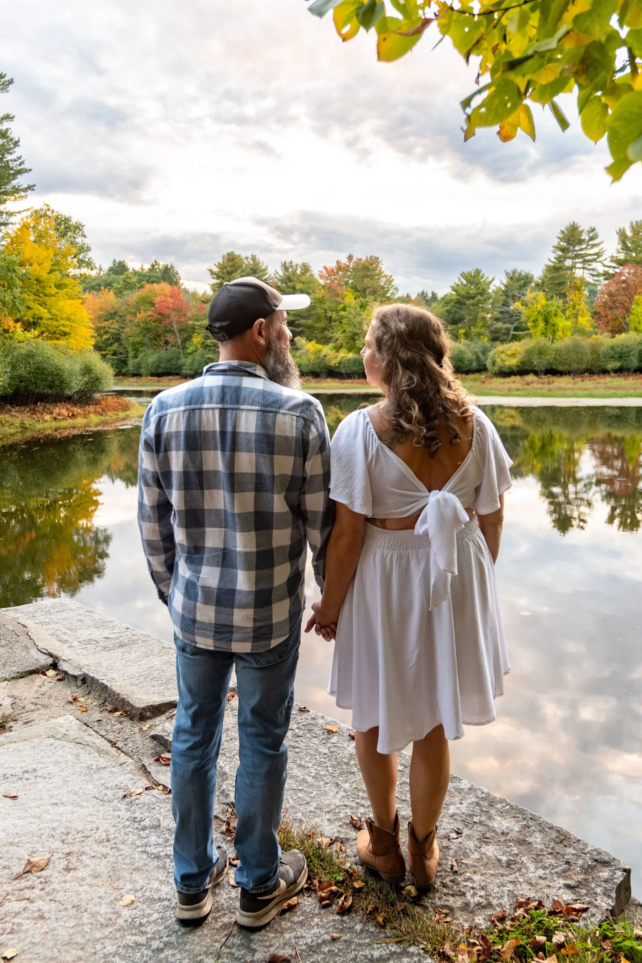 Alison and Dave from behind, holding hands and looking out over the pond with the bow of her dress showing at Vaughan Woods in Hallowell Maine fall engagement photography VMG Productions