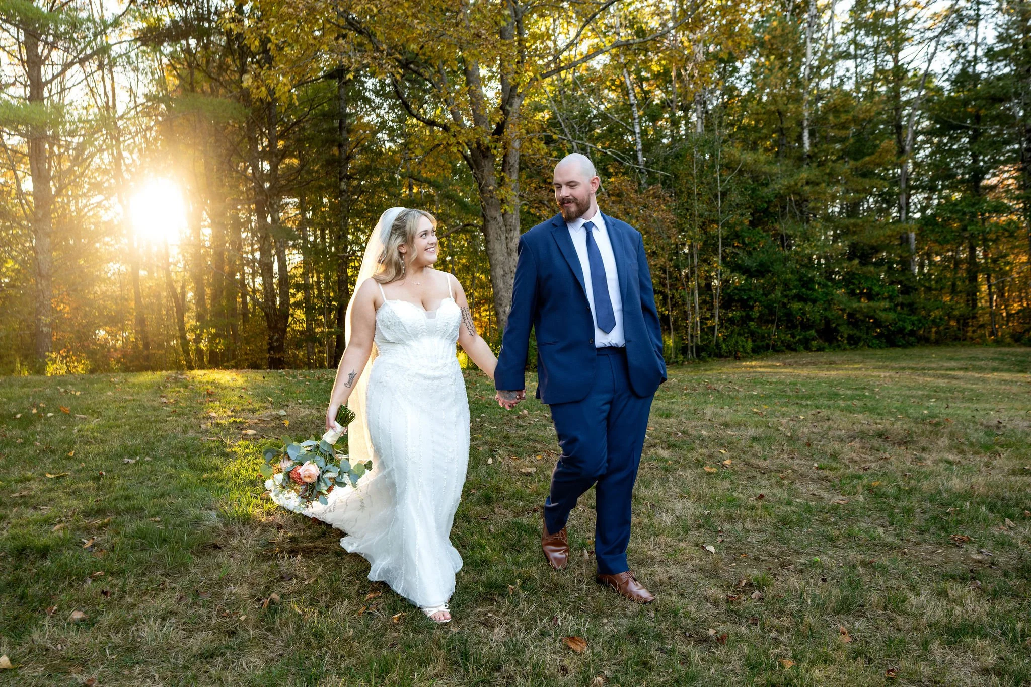 Ellie and Sam walking hand in hand across the lawn with the sun low in the trees behind them at Boothbay Playhouse in Boothbay Maine fall wedding photography VMG Productions