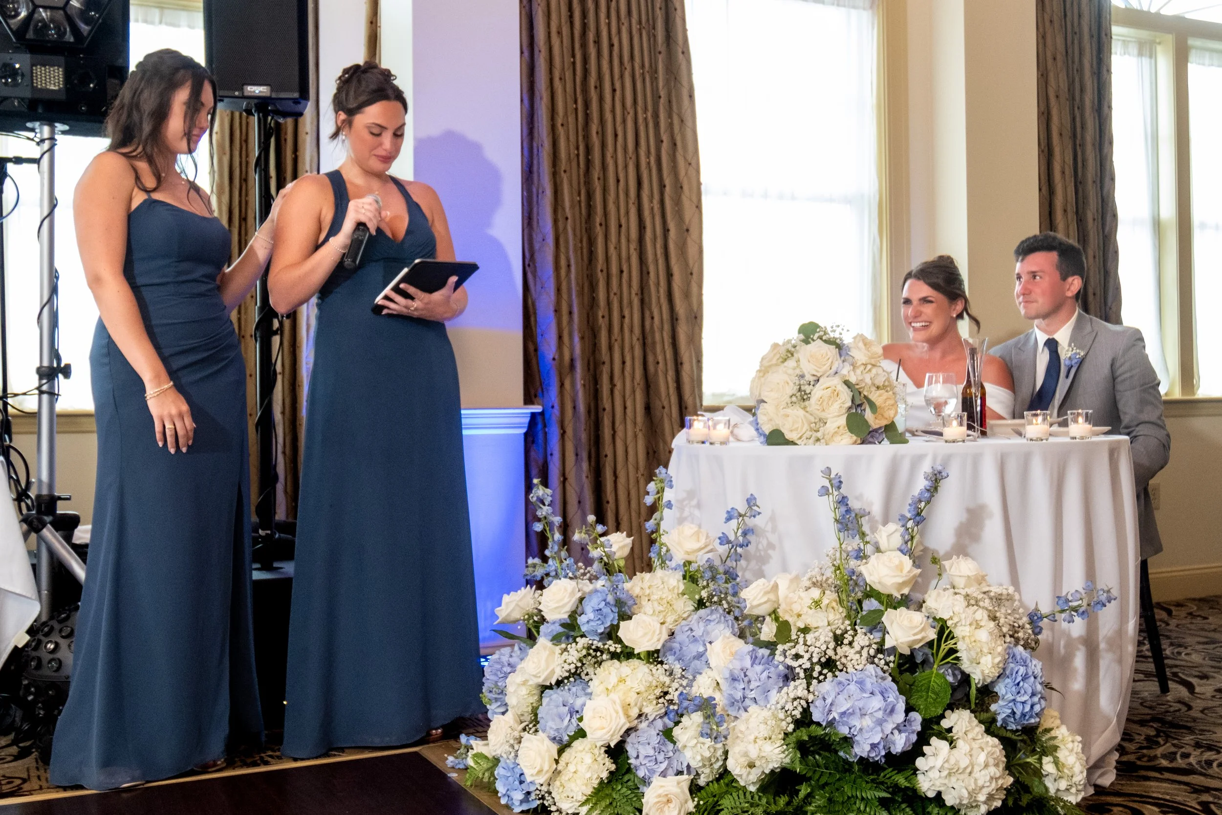 Caroline Pirolli's second maid of honor in navy reading her speech with her sister standing beside her for support as Caroline laughs from the sweetheart table at Union Bluff Hotel in York Maine wedding photography VMG Productions