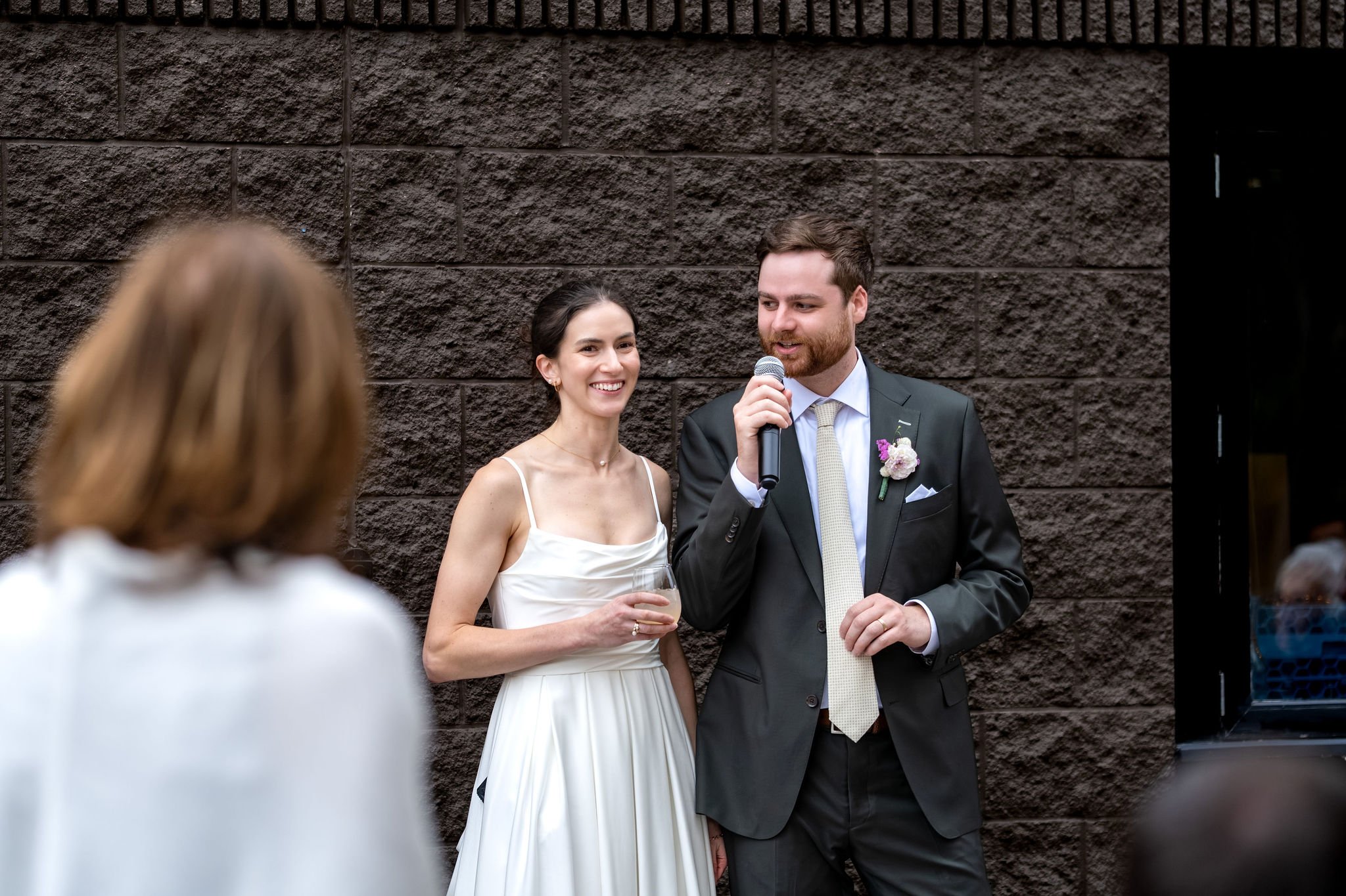 Jack thanking everyone into the microphone with Victoria smiling beside him on the patio at The Cellars at Allagash Brewing Company in Portland Maine fall wedding photography VMG Productions