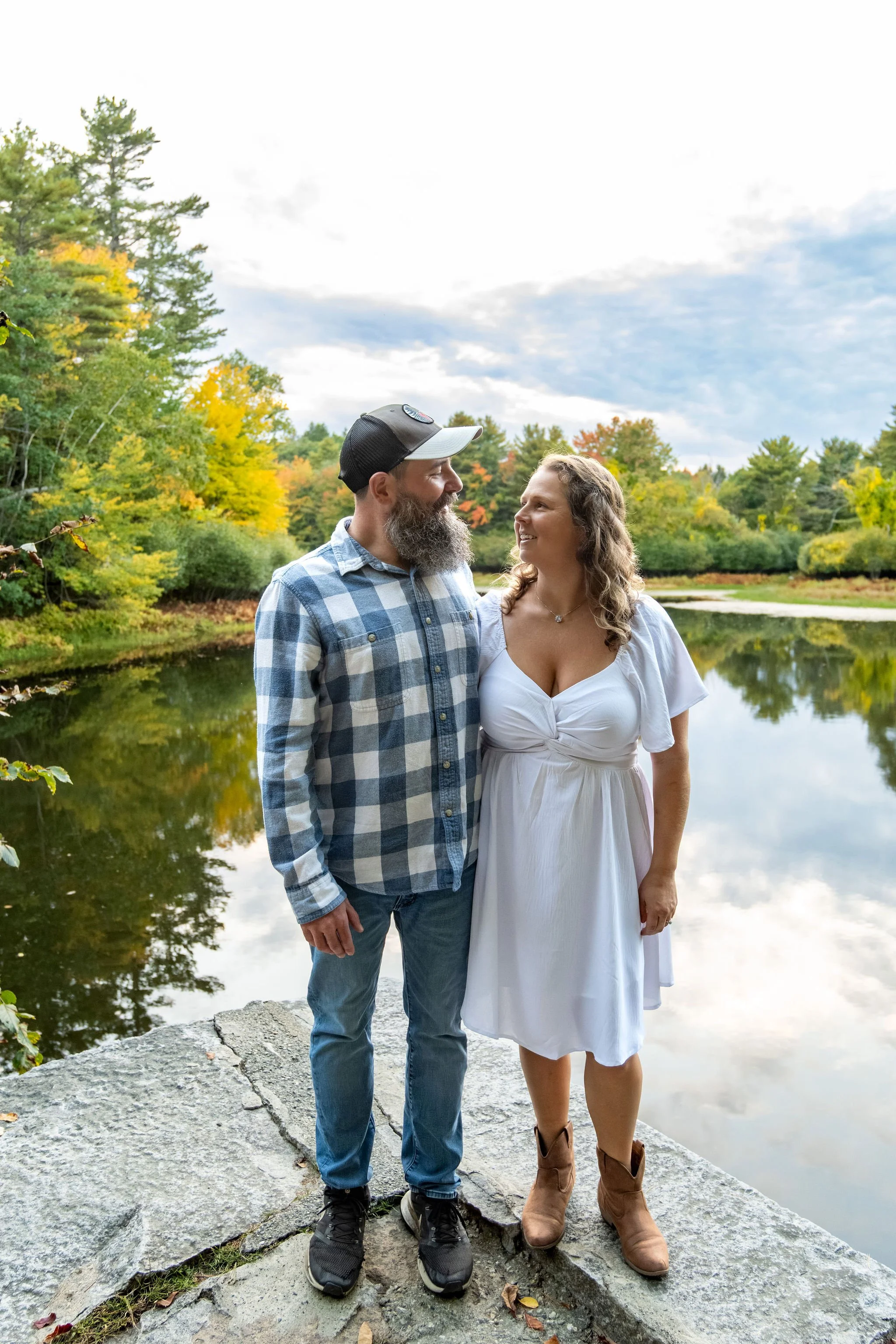 Alison and Dave looking at each other at the water's edge with the clouds reflected in the pond at Vaughan Woods in Hallowell Maine fall engagement photography VMG Productions