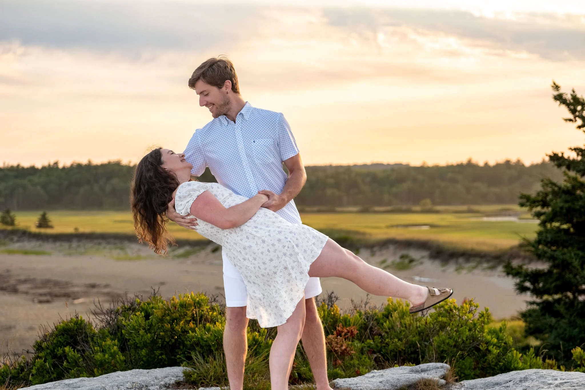 Lucas dipping Kirstie for a kiss on the headland above the beach at sunset during their engagement session at Reid State Park in Georgetown Maine engagement photography VMG Productions