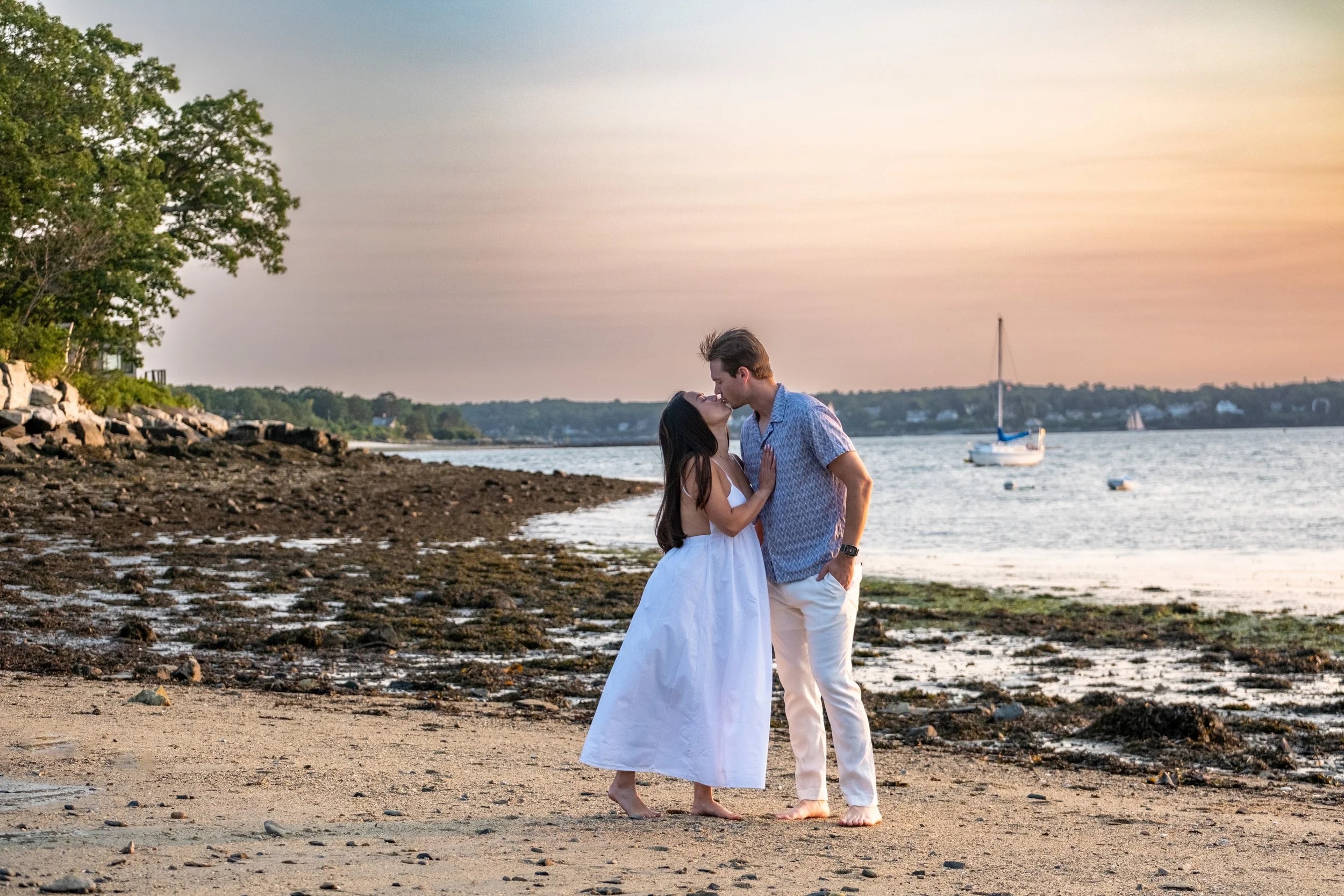 Couple kissing on the rocky beach with a sailboat behind, Peaks Island, Portland, Maine, engagement session by VMG Productions